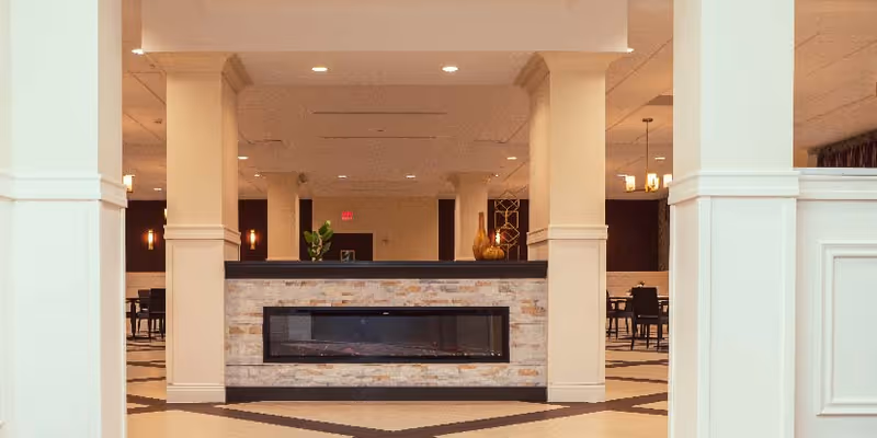 Interior view of a spacious common area with a modern electric fireplace set in a stone surround, flanked by large white columns. The room features a patterned floor and several dining tables and chairs in the background under warm lighting.