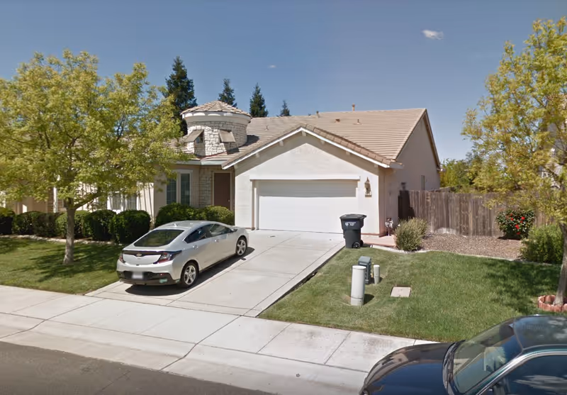 Front exterior view of a single-story house with a two-car garage, a silver car parked in the driveway, green lawn, trees, and a wooden fence on the right side under a clear blue sky.