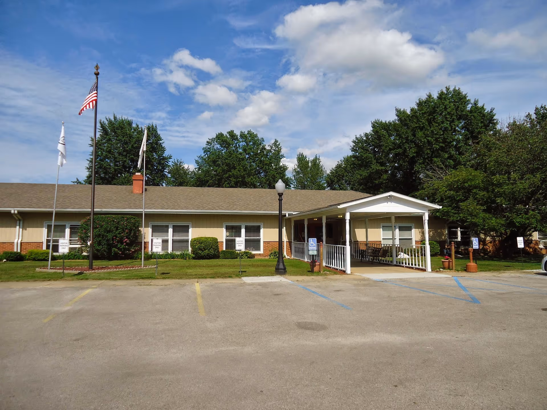 Exterior view of Casey Health Care Center, a single-story building with a beige and brick facade. There are three flagpoles with flags, a covered entrance with a wheelchair accessible ramp, and several parking spaces including handicapped spots. Trees and bushes surround the building under a partly cloudy sky.