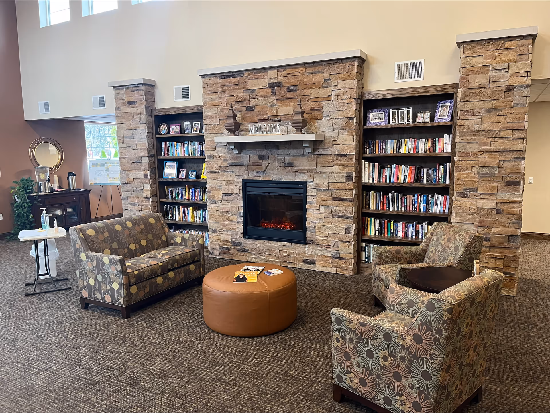 A cozy common area with a stone fireplace in the center, flanked by built-in bookshelves filled with books and framed photos. There are two patterned armchairs and a matching loveseat arranged around a round brown ottoman with magazines on top. The room has carpeted flooring and a warm, inviting atmosphere.