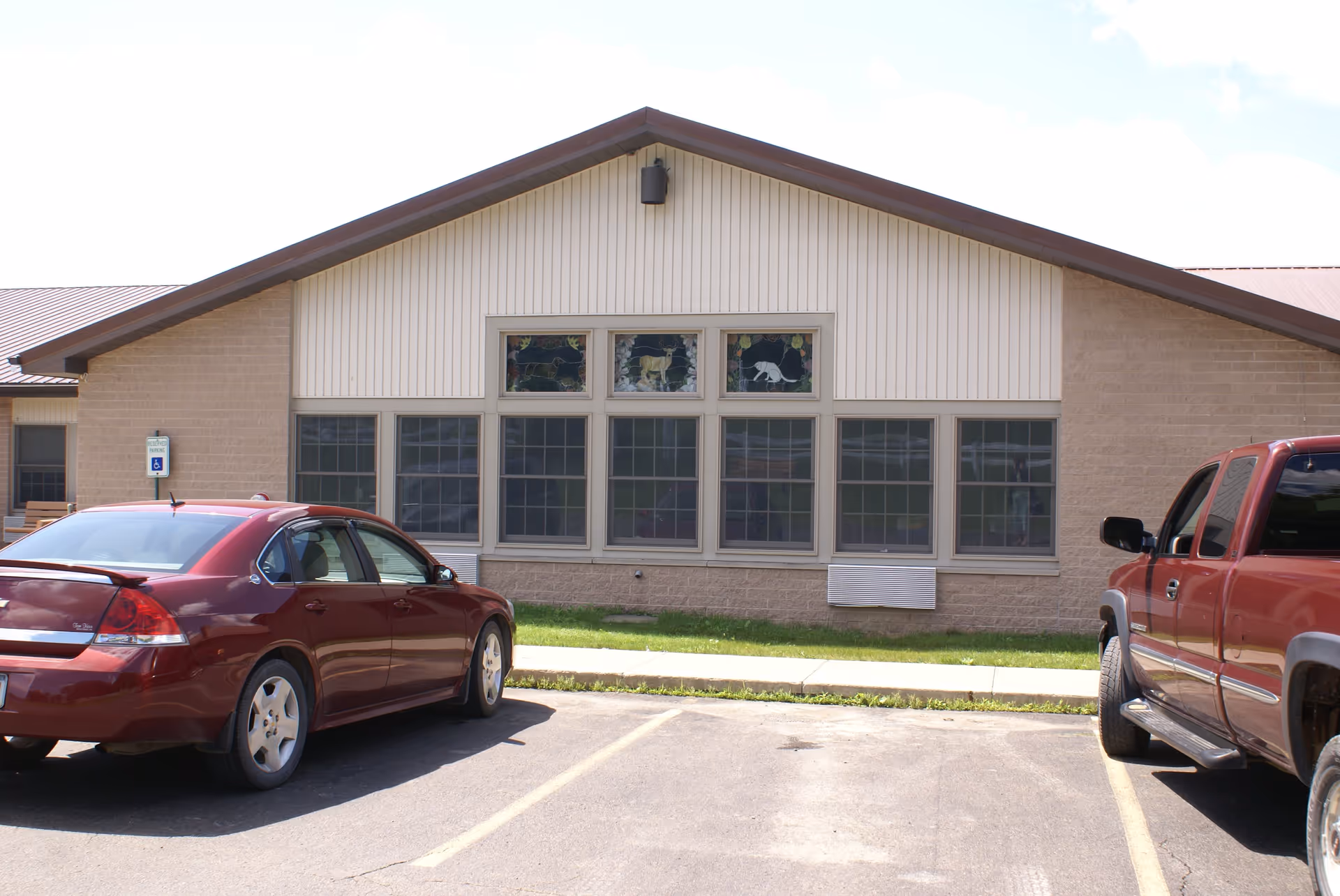Exterior view of a single-story building with beige siding and a brown roof. There are large windows with decorative stained glass panels above them, depicting animals. Two red vehicles are parked in front of the building, one sedan and one pickup truck. A reserved parking sign for handicapped parking is visible near the left side of the building.