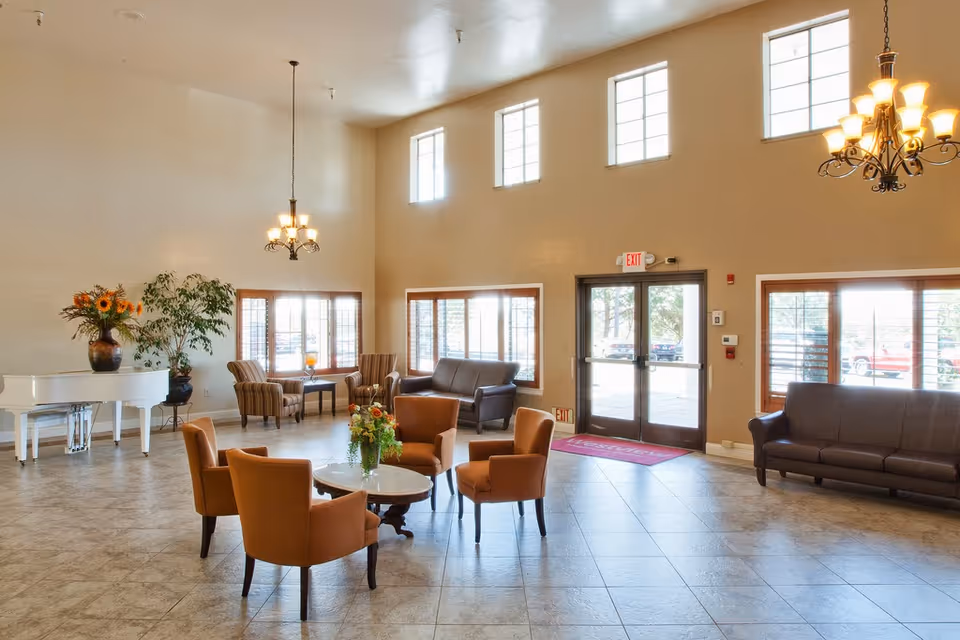A spacious and well-lit common area with high ceilings and large windows. The room features a white grand piano with a vase of sunflowers, several comfortable chairs arranged around a small round table with a floral centerpiece, and additional seating including armchairs and sofas near the windows. Two chandeliers hang from the ceiling, and double glass doors lead outside.