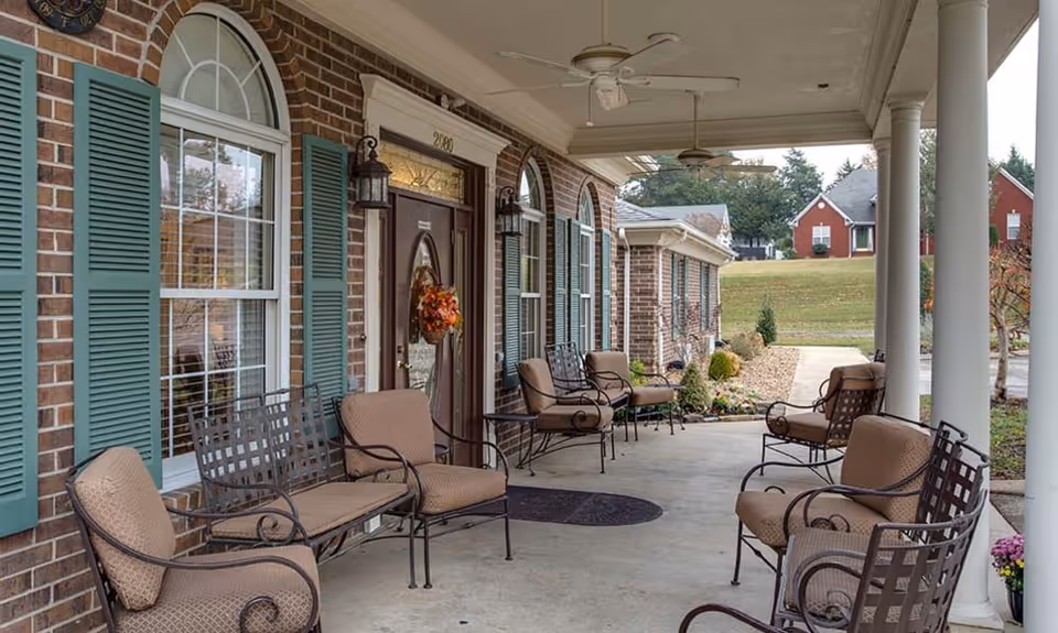 Covered front porch of a brick senior living building with cushioned metal chairs, white columns, and a wreath on the door.