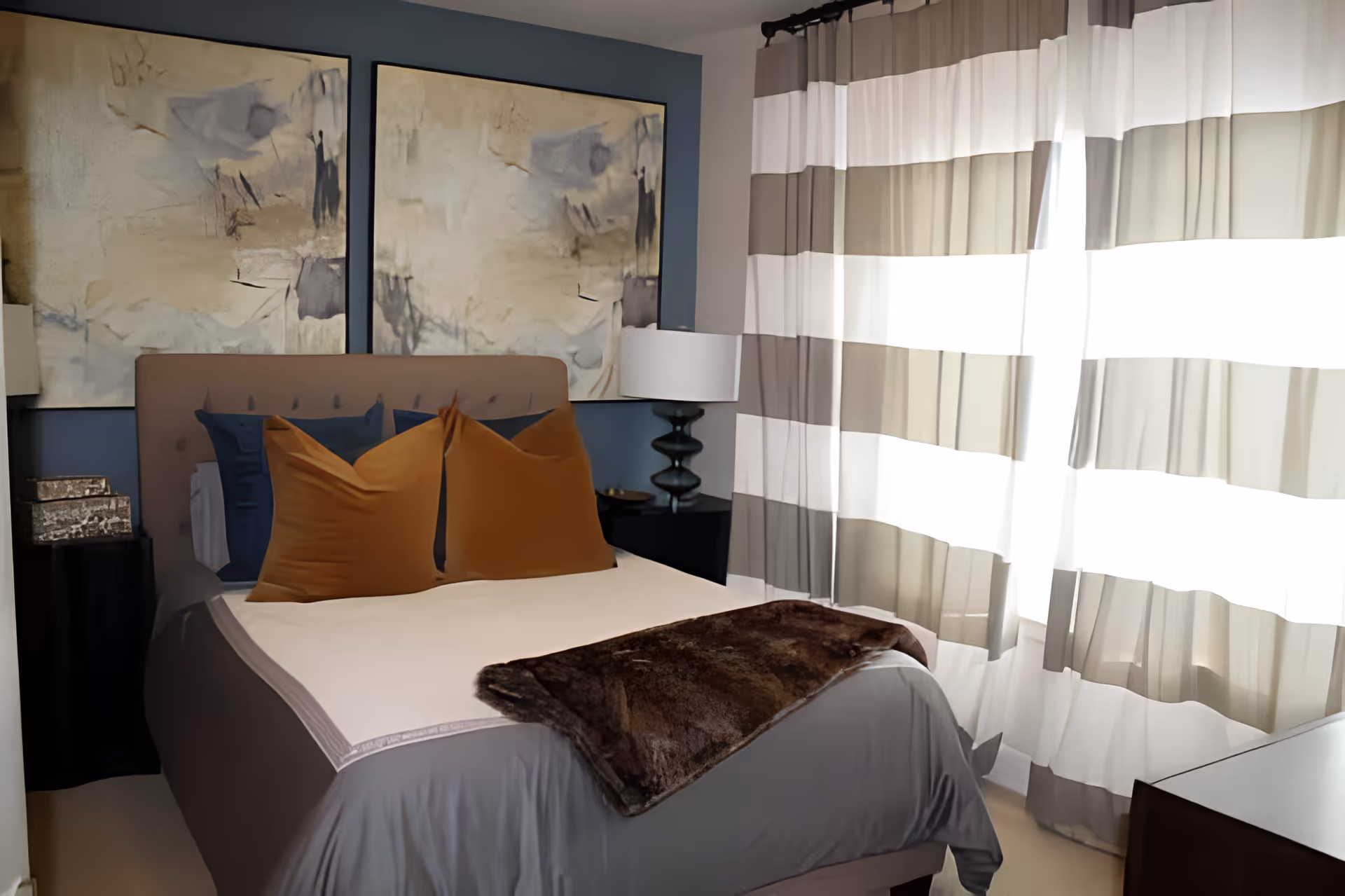 A cozy bedroom with a neatly made bed featuring brown and blue pillows, a brown throw blanket, and a beige headboard. Two abstract paintings hang on the blue wall behind the bed. A bedside table with a lamp is visible on the right side, and large windows with striped curtains allow natural light to fill the room.