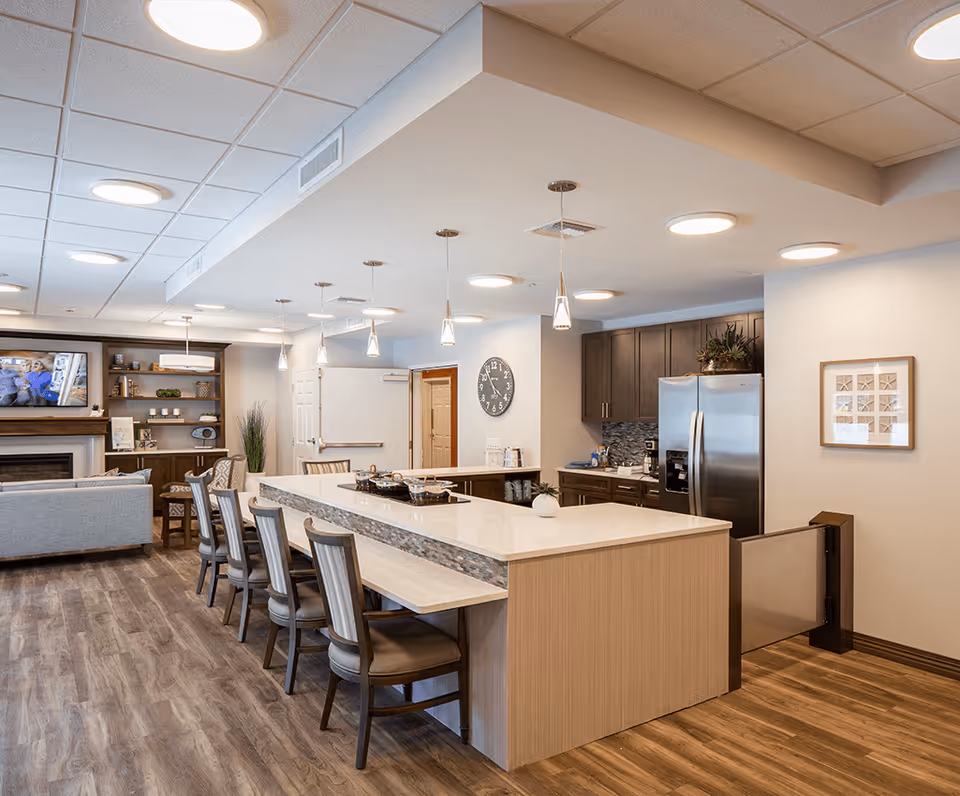 A modern, open-concept kitchen and living area in a senior living facility. The kitchen features a large island with seating for six, pendant lighting, stainless steel refrigerator, and dark wood cabinets. The living area includes a fireplace, a mounted TV, and a gray sofa. The space has wood flooring and neutral-colored walls with decorative elements.