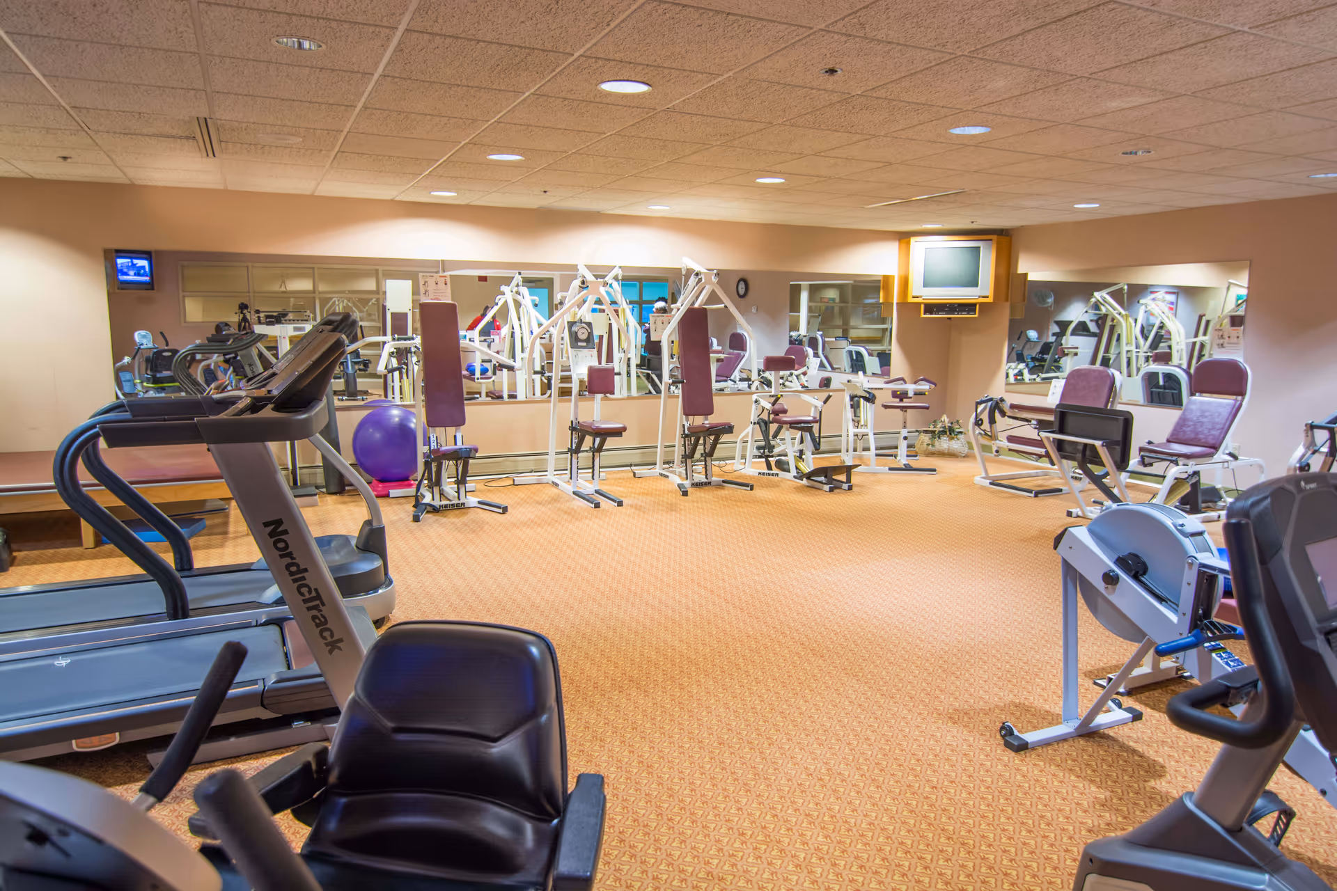 A fitness room with various exercise equipment including treadmills, weight machines, a rowing machine, and an exercise ball. The room has beige walls, a patterned carpet, and large mirrors on the walls. A small television is mounted on the wall.