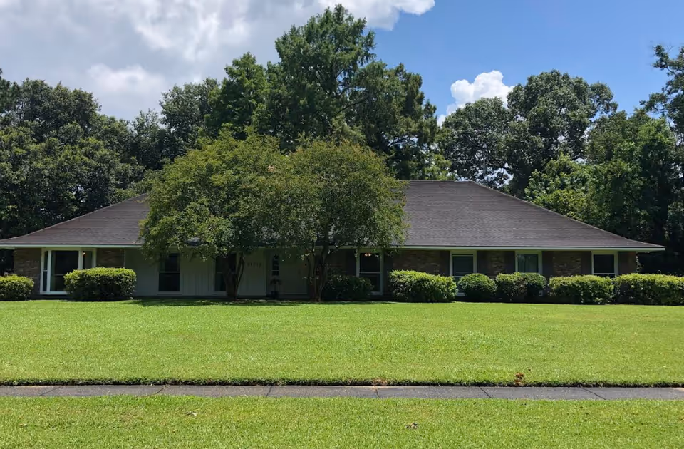 Single-story residential building with a wide front lawn, surrounded by trees under a partly cloudy sky.