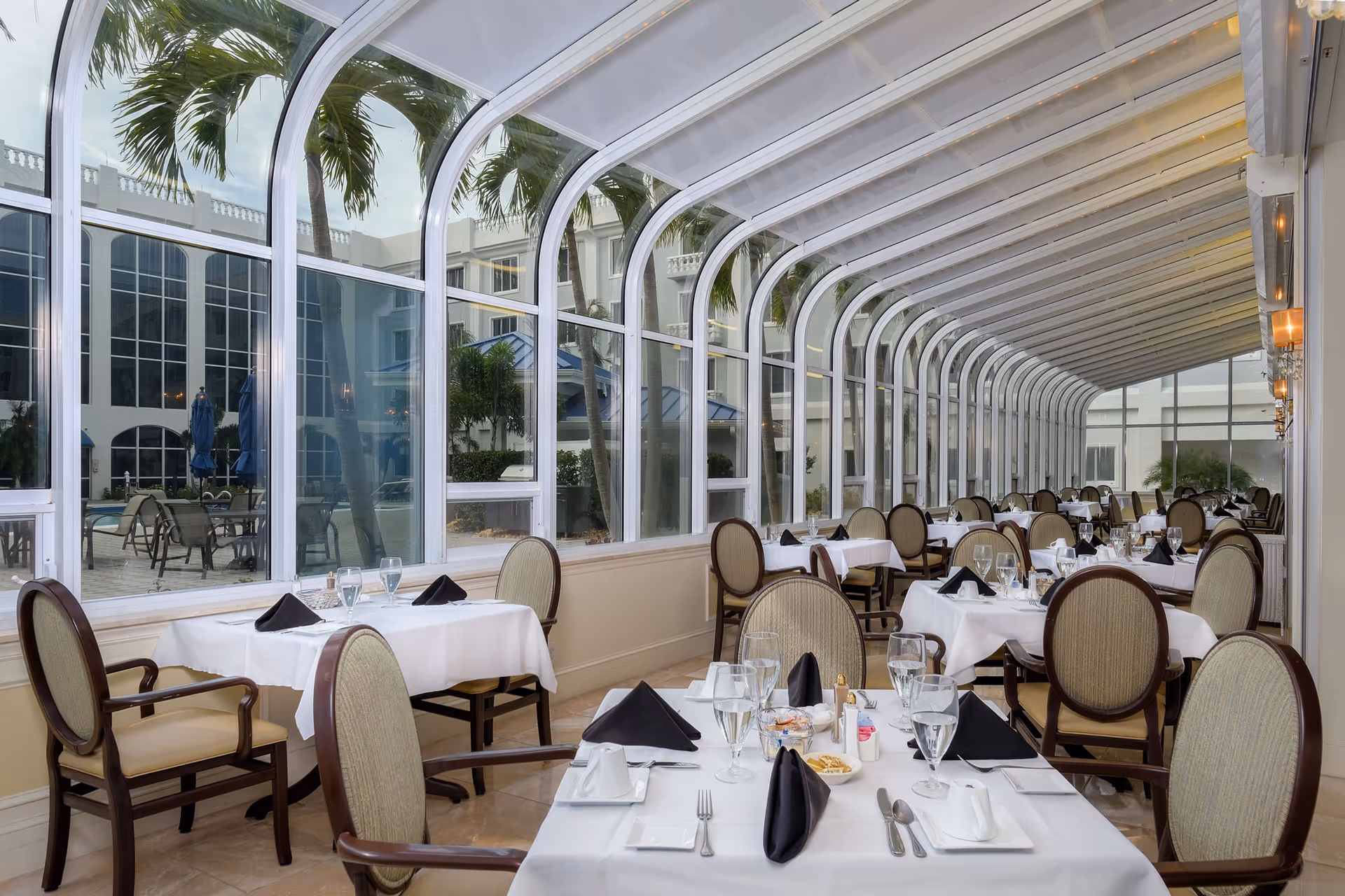 Sunlit glass-enclosed dining room with tables set with white tablecloths, black napkins, and chairs overlooking an outdoor courtyard and pool.