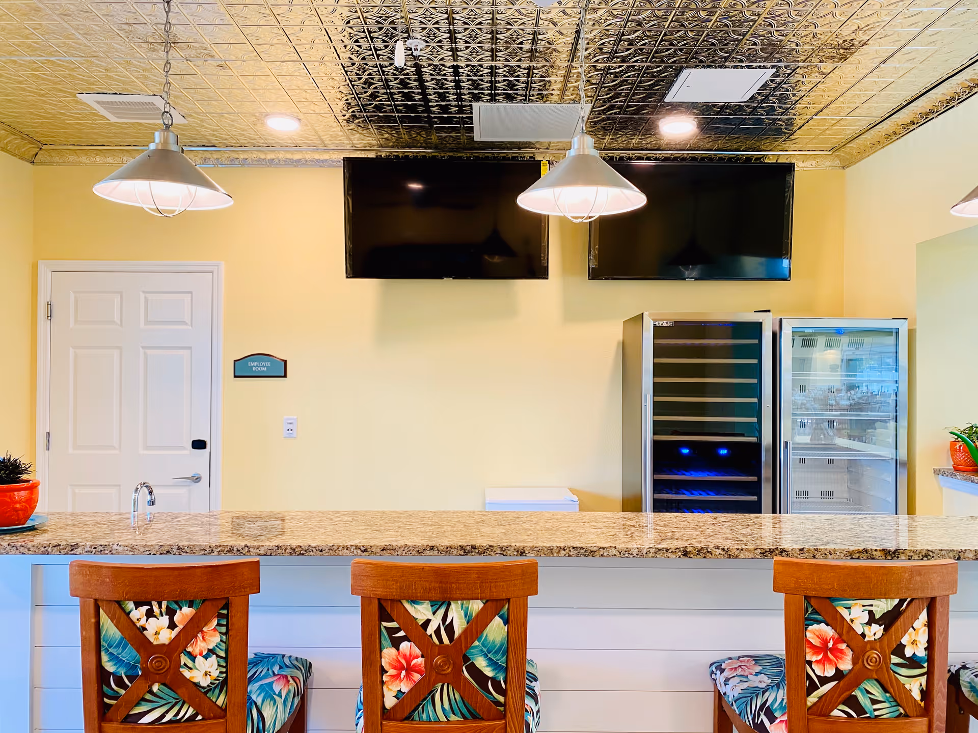 Interior view of a facility counter area with a granite countertop and three wooden chairs with floral patterned cushions. Behind the counter, there are two large flat-screen TVs mounted on a pale yellow wall, a door labeled 'Employee Room', and two glass-front refrigerators. The ceiling has a decorative metallic pattern with hanging pendant lights.
