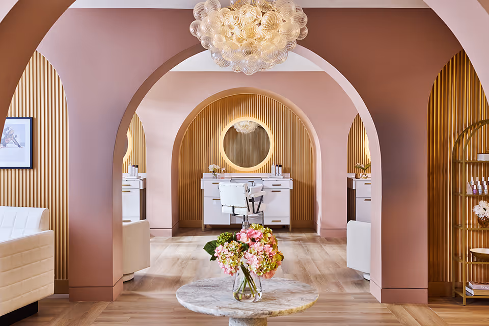 A stylish interior space featuring soft pink arched walls and wooden slatted panels. The room includes a round marble table with a vase of pink and green flowers in the foreground, white cushioned chairs, a white vanity with a round mirror, and a modern chandelier hanging from the ceiling.