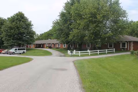 Brick senior care building with curved driveway, white fence, and green lawn.