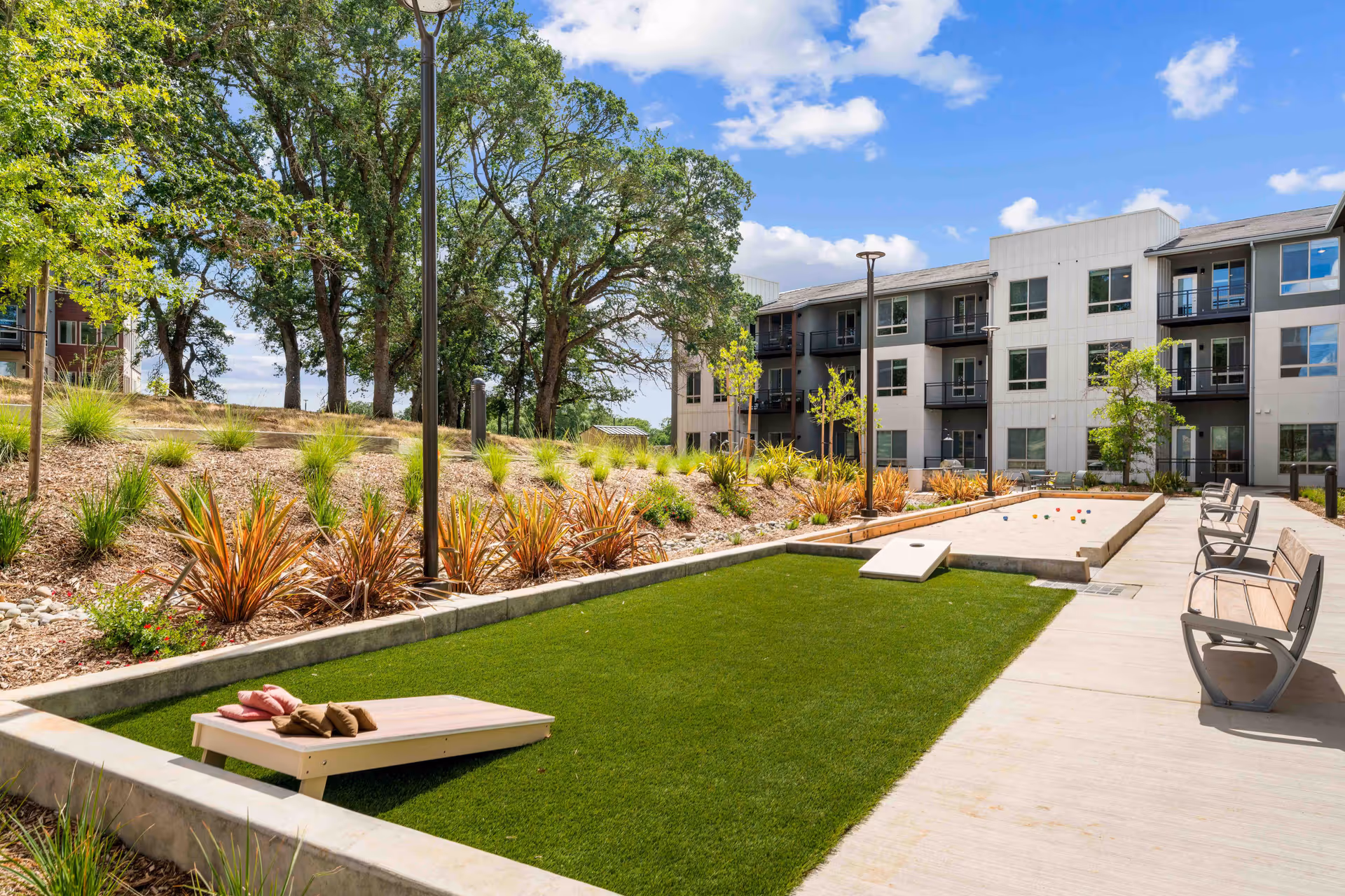 Outdoor recreational area at Revel Folsom featuring a bocce ball court and a cornhole game set on artificial grass, surrounded by benches and landscaping with trees and shrubs, with a multi-story residential building in the background under a partly cloudy sky.