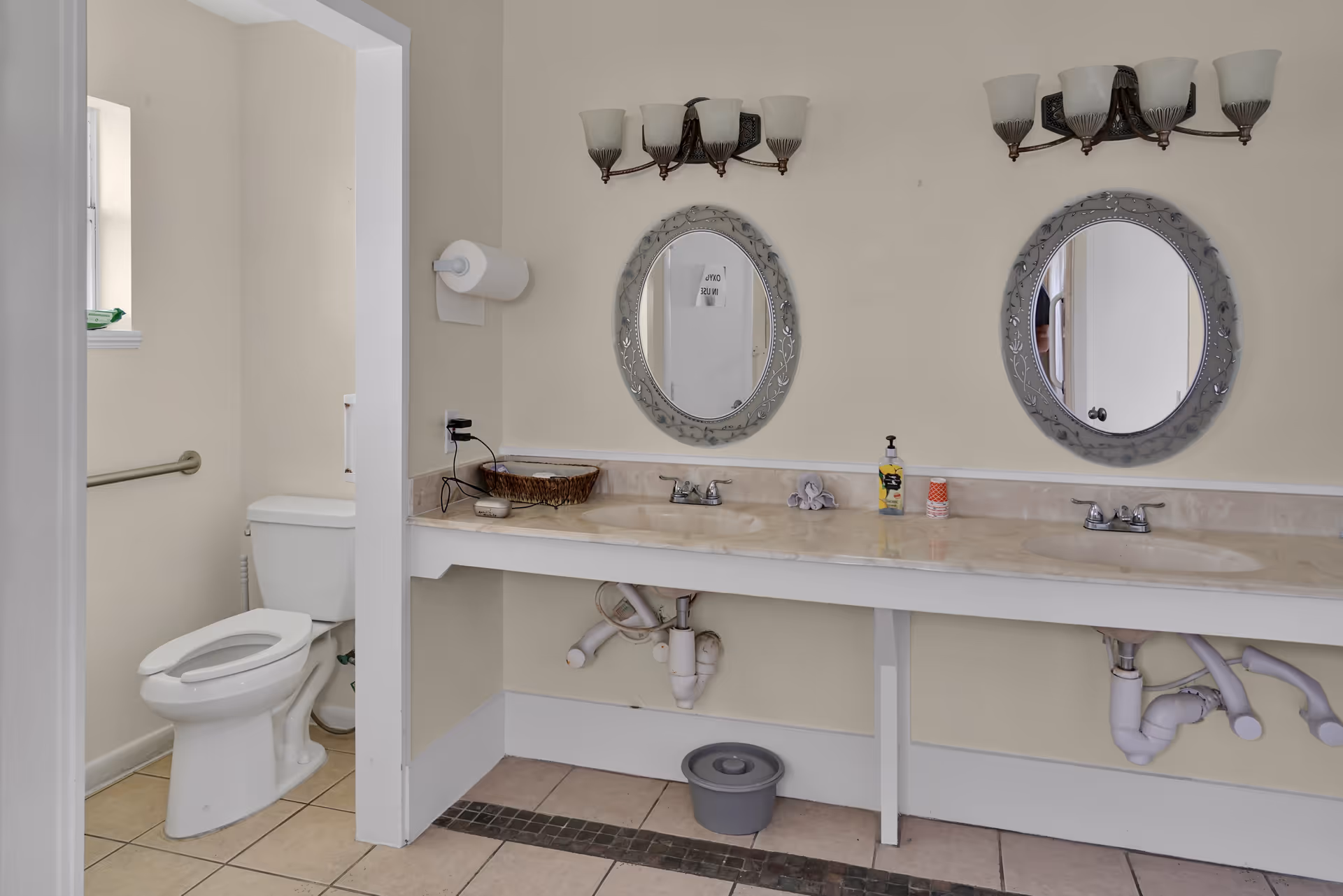 Bathroom with a double-sink vanity, two oval mirrors and wall light fixtures, and a toilet visible in a side stall.