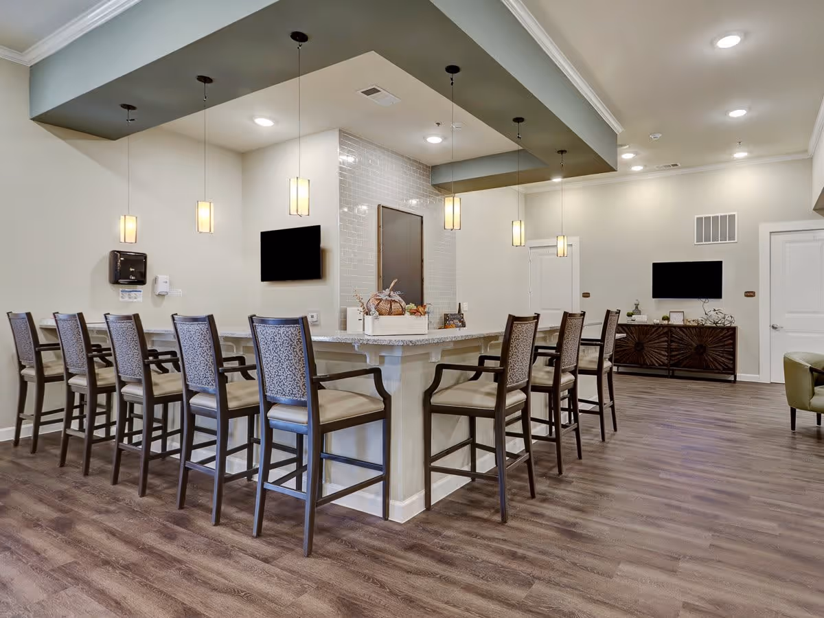 Interior view of a communal dining or bar area in The Lodge at Pine Creek featuring a long counter with high chairs, pendant lights hanging from the ceiling, two wall-mounted televisions, and a decorative centerpiece on the counter. The room has wood flooring and light-colored walls with modern decor.