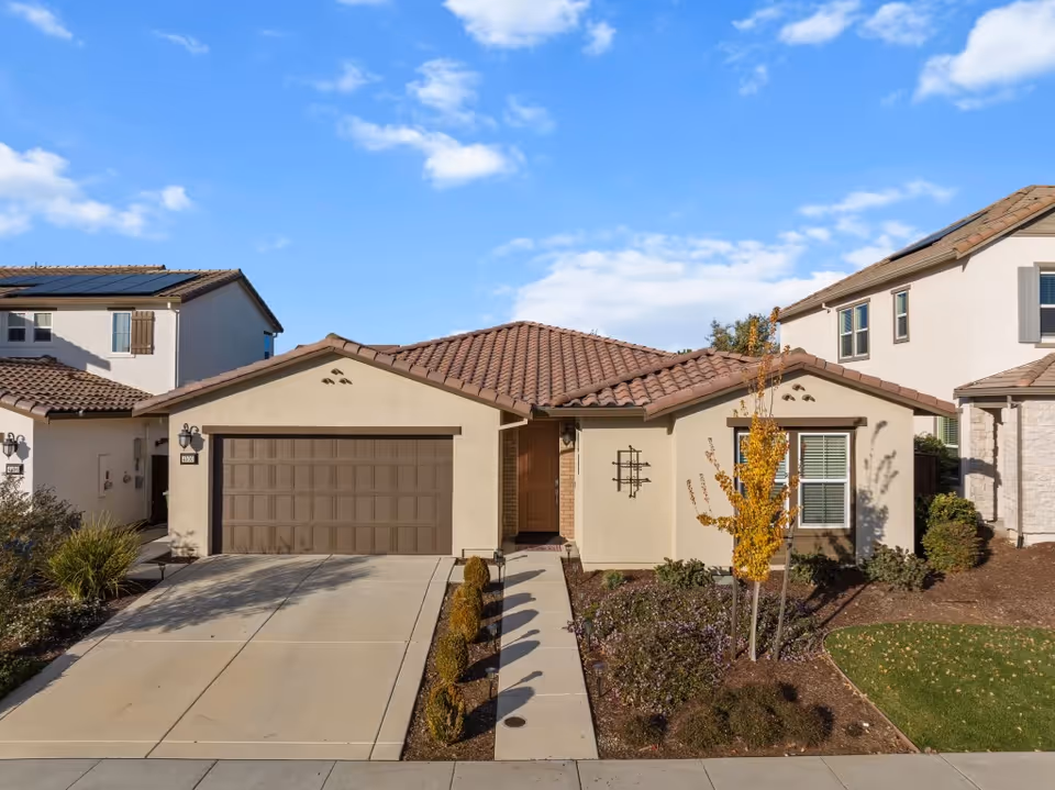 Single-story stucco home with a two-car garage, tiled roof, driveway and small landscaped front yard under a blue sky.