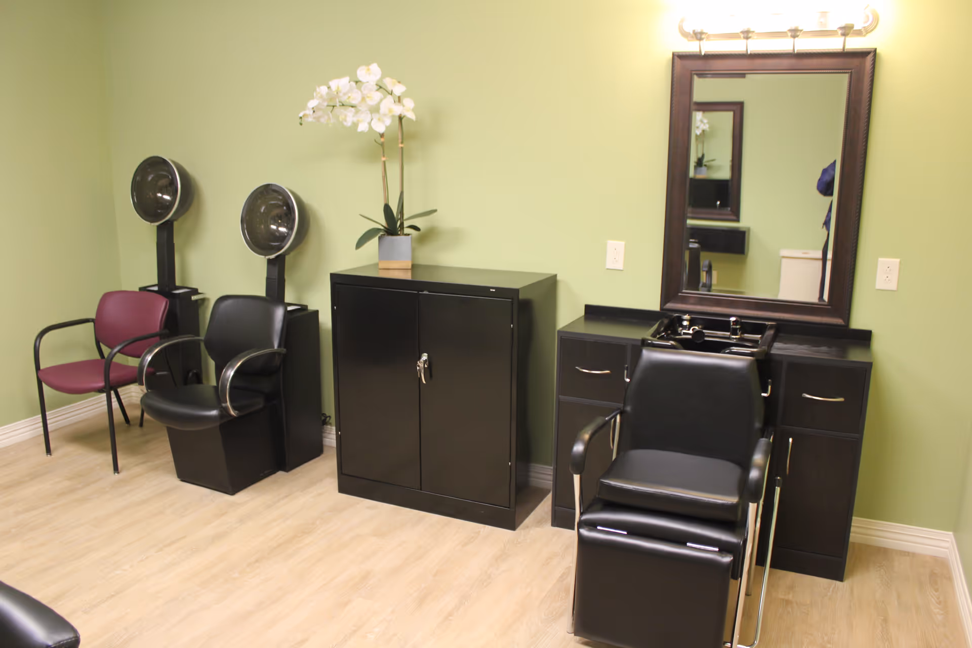 Small salon area with hair-washing chairs, hooded dryers, cabinets, and a mirror against a green wall.