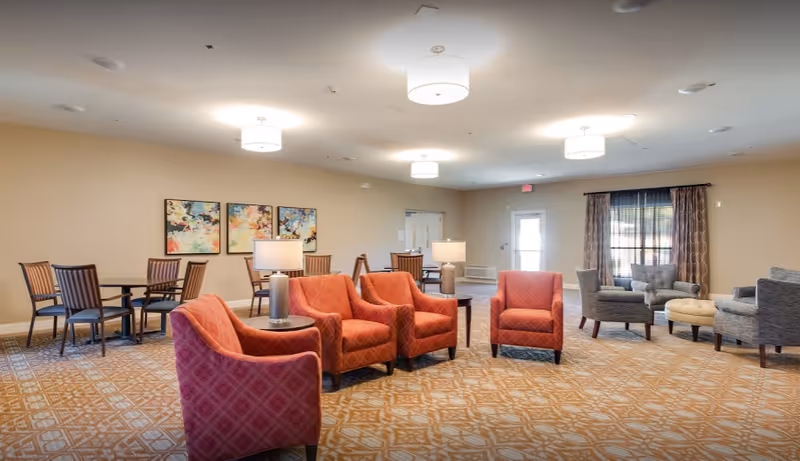 A spacious common area in an assisted living facility featuring multiple seating arrangements including red upholstered armchairs, gray armchairs with a round ottoman, and wooden dining tables with chairs. The room has beige walls, patterned carpet, ceiling lights, and abstract artwork on the wall.
