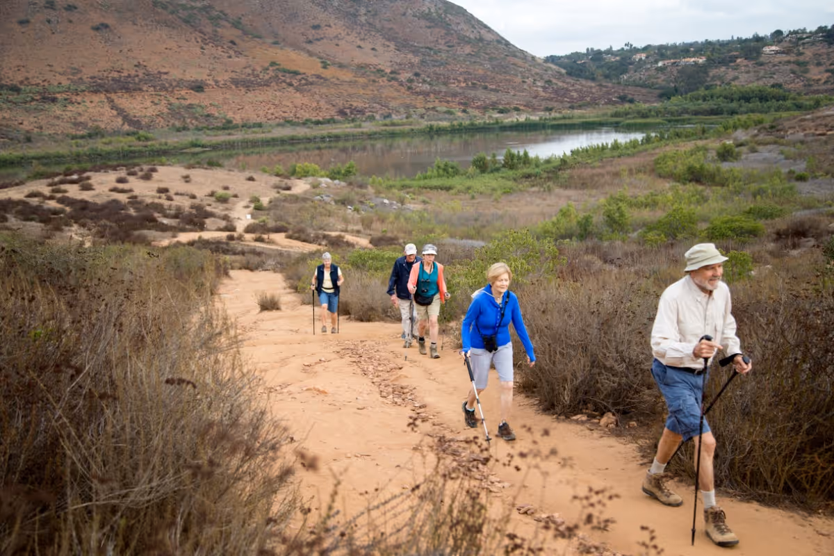 A group of five elderly people hiking on a dirt trail in a dry, hilly landscape with sparse vegetation and a body of water in the background. They are using walking sticks and dressed in outdoor hiking attire.