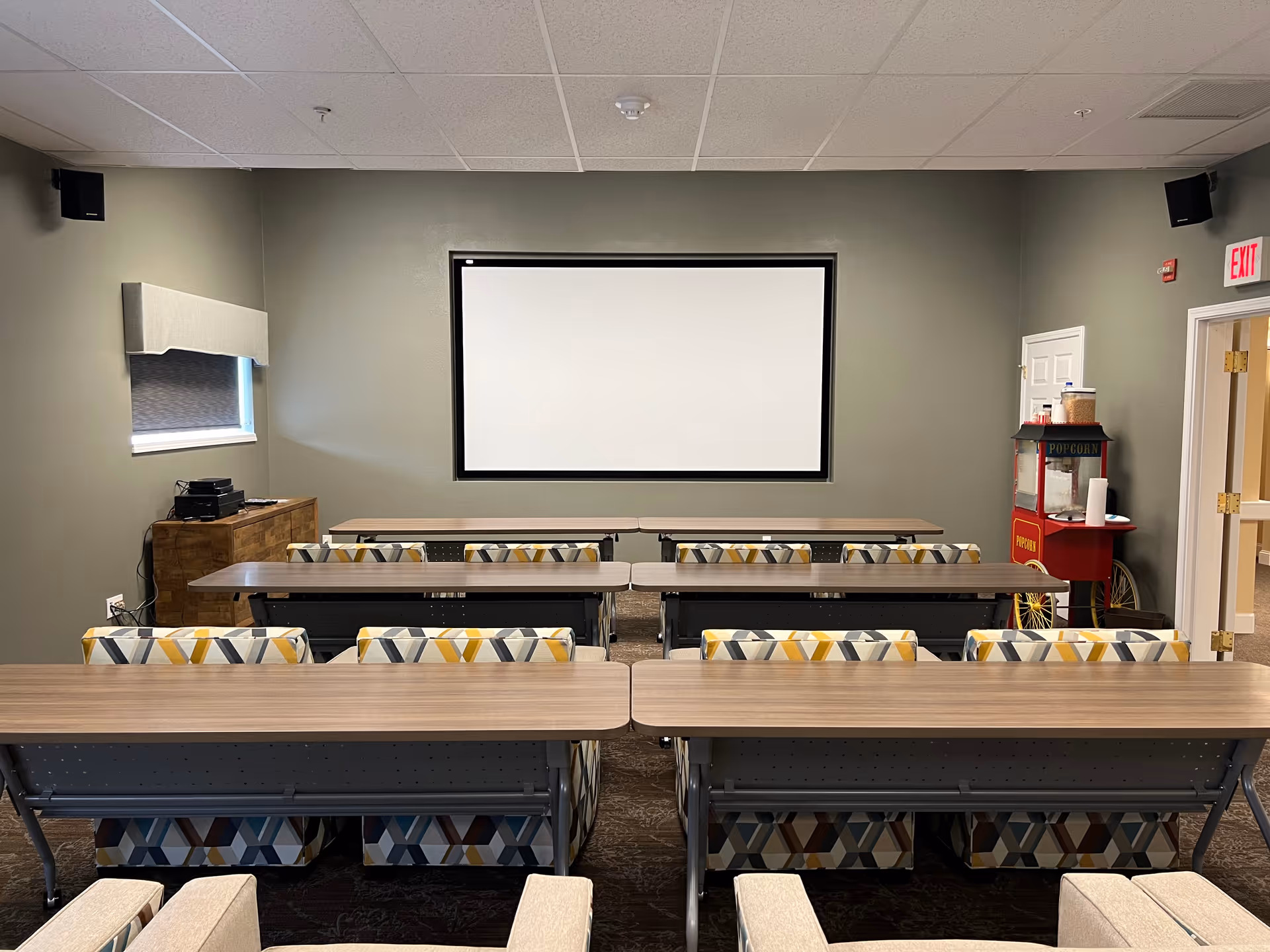 Small screening/classroom with rows of tables and patterned chairs facing a large blank projection screen and a popcorn machine on the right.