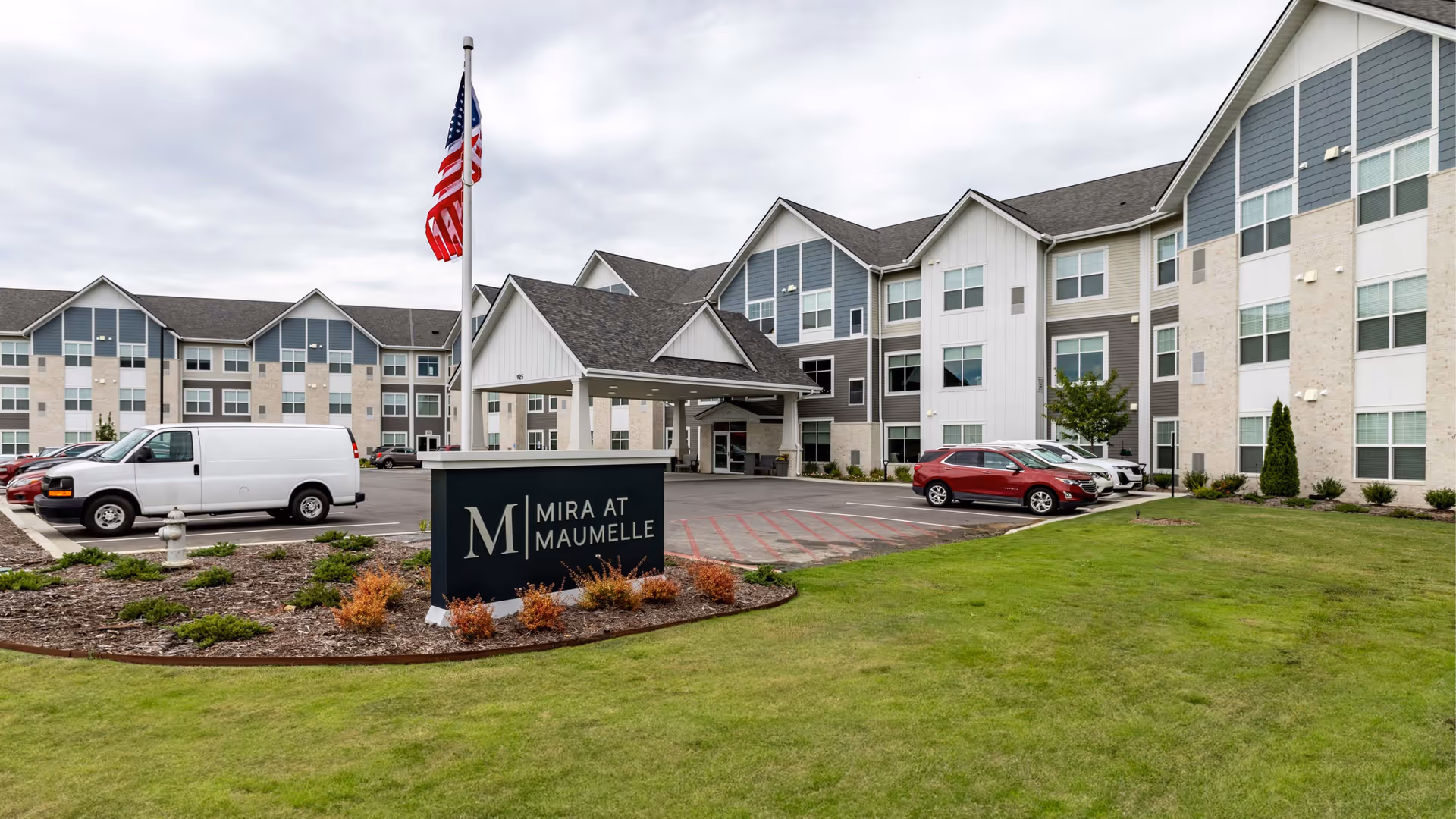 Front exterior of the Mira at Maumelle senior living building with an entrance canopy, sign, American flag, parked cars, and landscaped lawn.