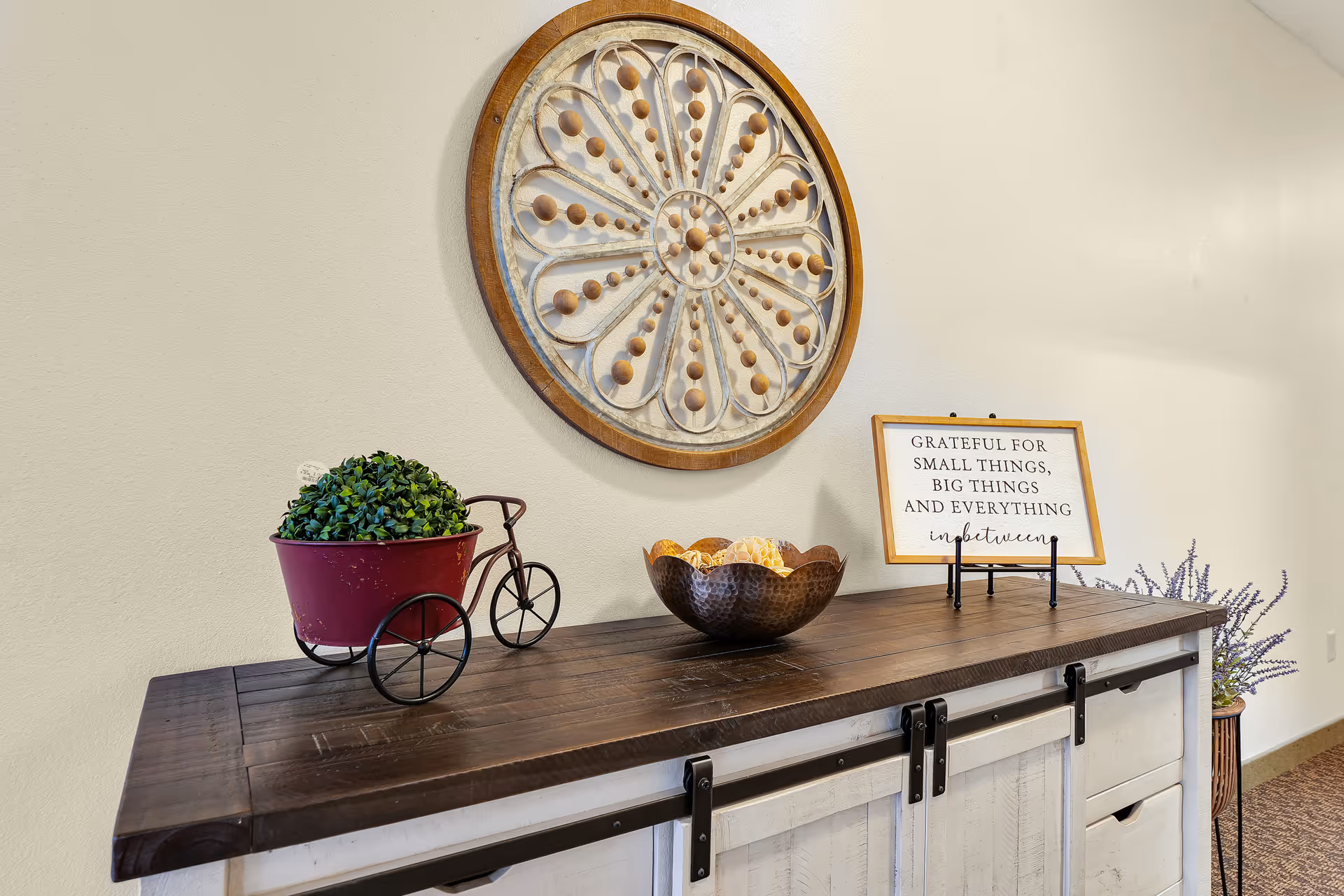 A decorative wooden sideboard with a dark wood top and white sliding barn doors. On top of the sideboard is a small red metal tricycle planter with a green plant, a hammered metal bowl with decorative balls, and a framed sign that reads 'GRATEFUL FOR SMALL THINGS, BIG THINGS AND EVERYTHING inbetween'. Above the sideboard hangs a large round wooden wall decoration with a floral pattern made of wooden beads. The wall and carpeted floor are visible in the background.