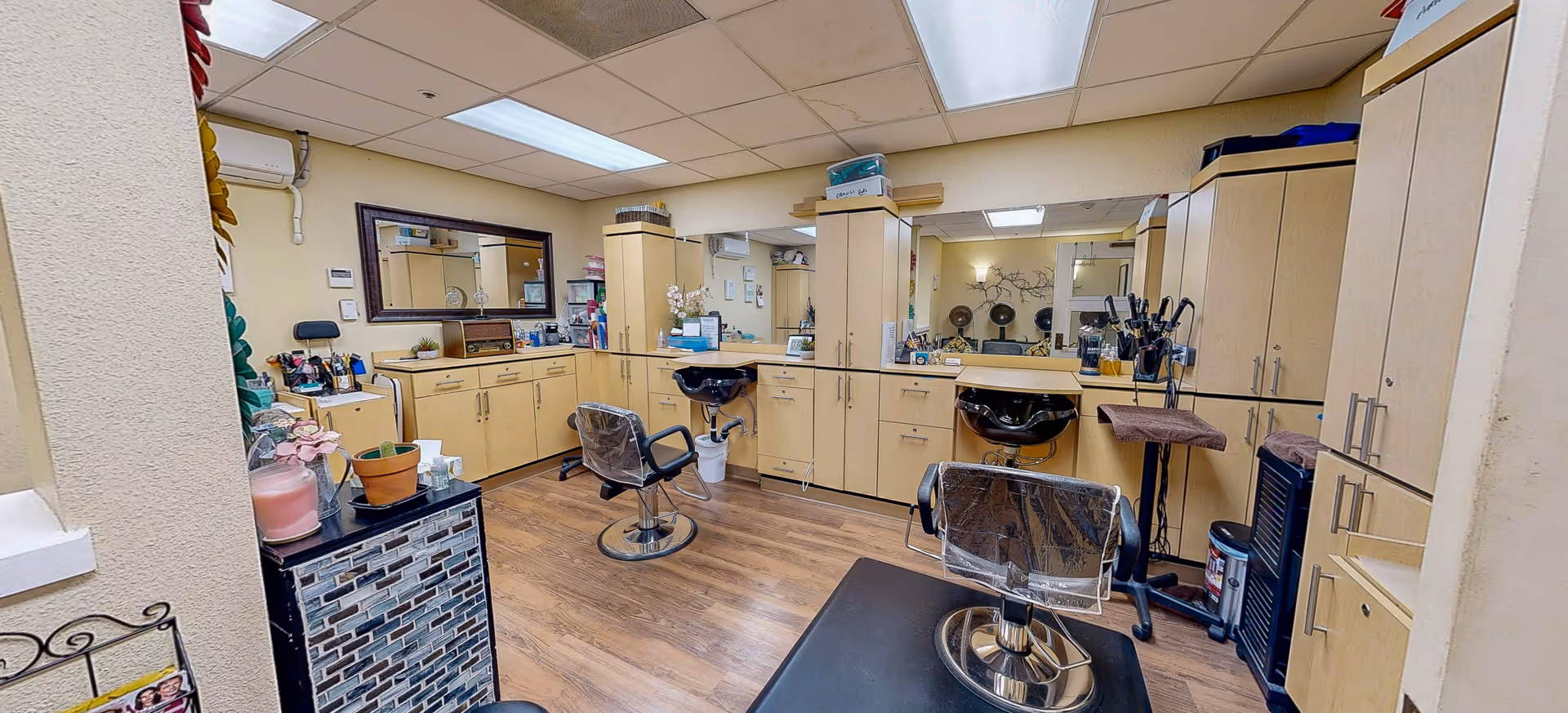 Interior view of a senior living facility's hair salon with two salon chairs covered in plastic, two black hair washing sinks, light wood cabinetry, large mirrors, and various hair styling tools and products on the counters.