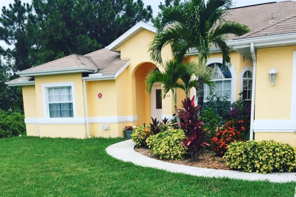 Exterior view of a single-story yellow building with white trim, a curved concrete pathway leading to the entrance, and landscaped garden beds with various plants and palm trees in front.