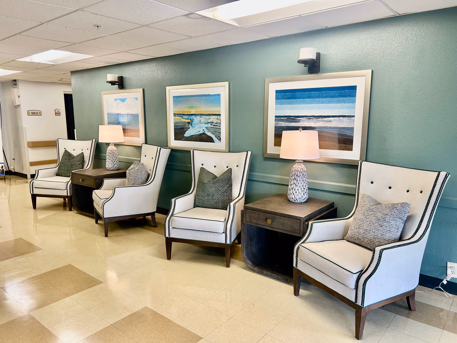 A seating area in a nursing and rehabilitation center with four white armchairs, each with a decorative pillow, arranged along a teal wall. Between the chairs are two dark wooden side tables, each holding a table lamp with a white patterned base and a white lampshade. Above the chairs are three framed landscape paintings featuring ocean and beach scenes. The floor is tiled in a beige and light brown checkerboard pattern.