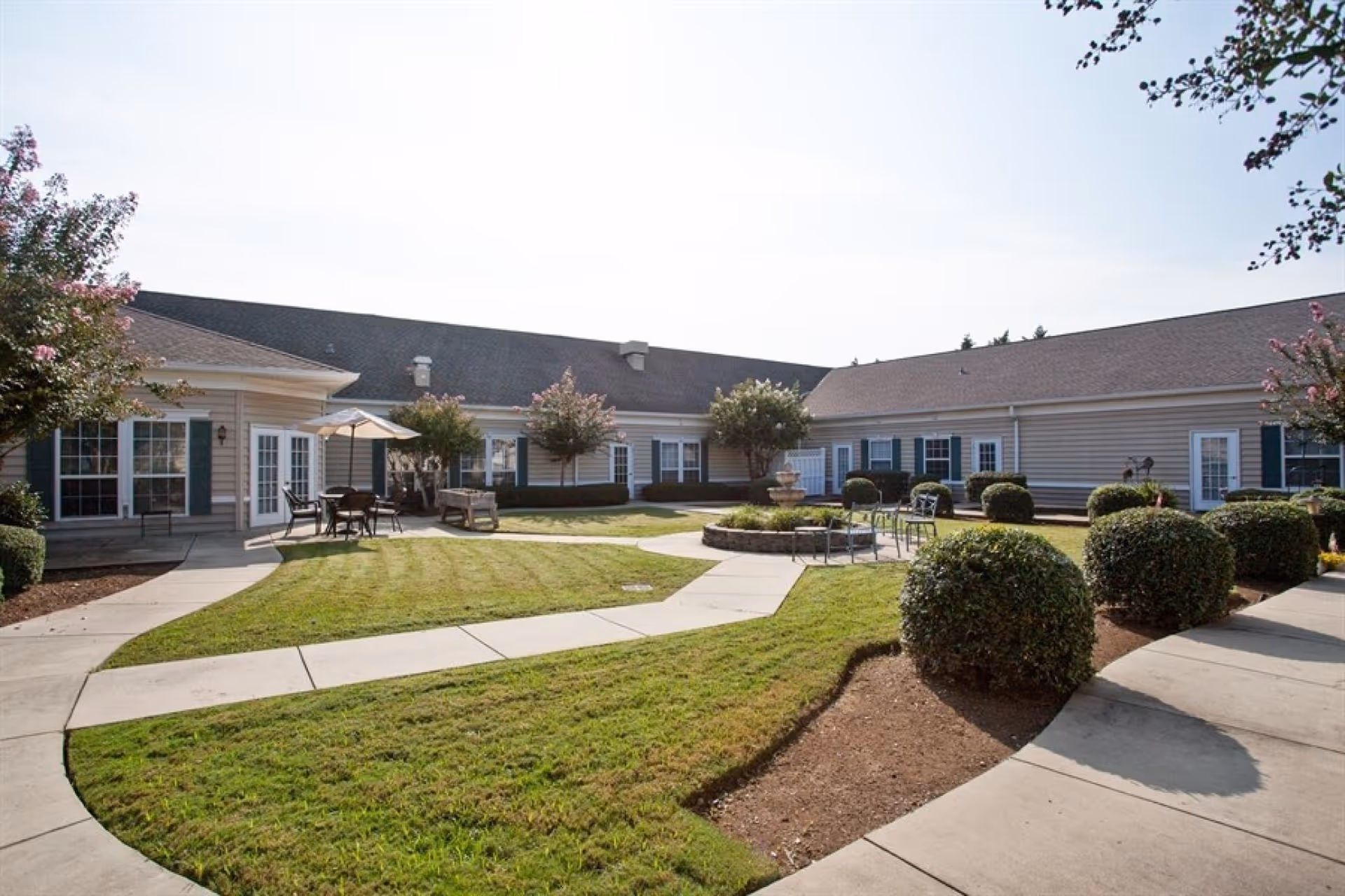 Sunlit courtyard with paved walkways, lawn, seating areas, shrubs, and single-story facility buildings surrounding it.