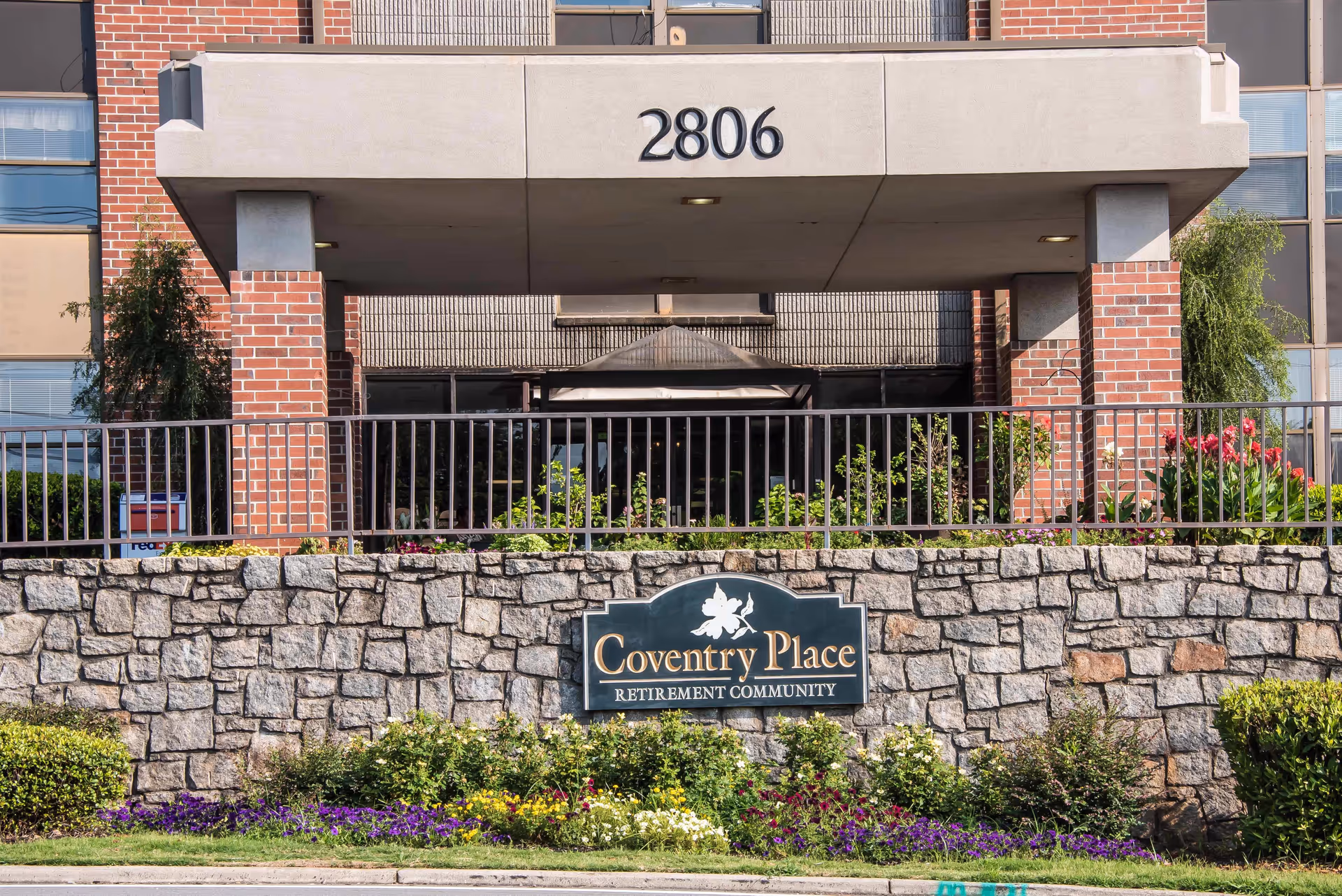 Entrance of Coventry Place retirement community with a stone wall and a sign displaying the community name. The building entrance is visible behind the wall with brick pillars and the number 2806 above the entrance.