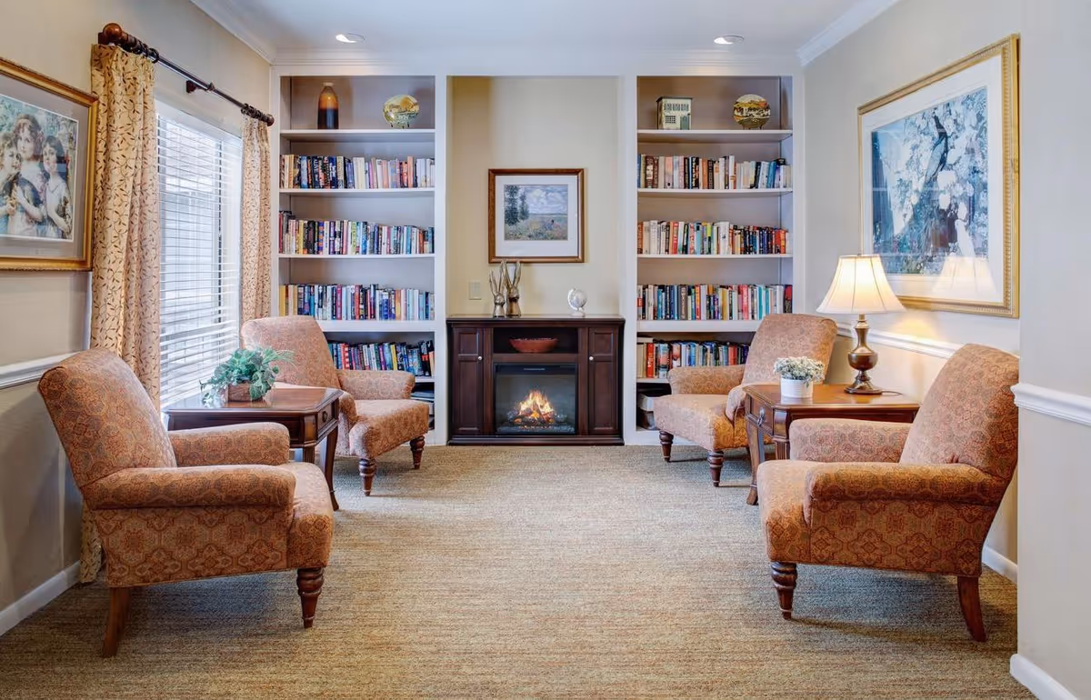 A cozy living room with four patterned armchairs arranged around two wooden side tables. A fireplace with a fire burning is centered between two built-in bookshelves filled with books. The room has beige walls, carpeted floor, framed artwork on the walls, a table lamp, and a window with curtains letting in natural light.