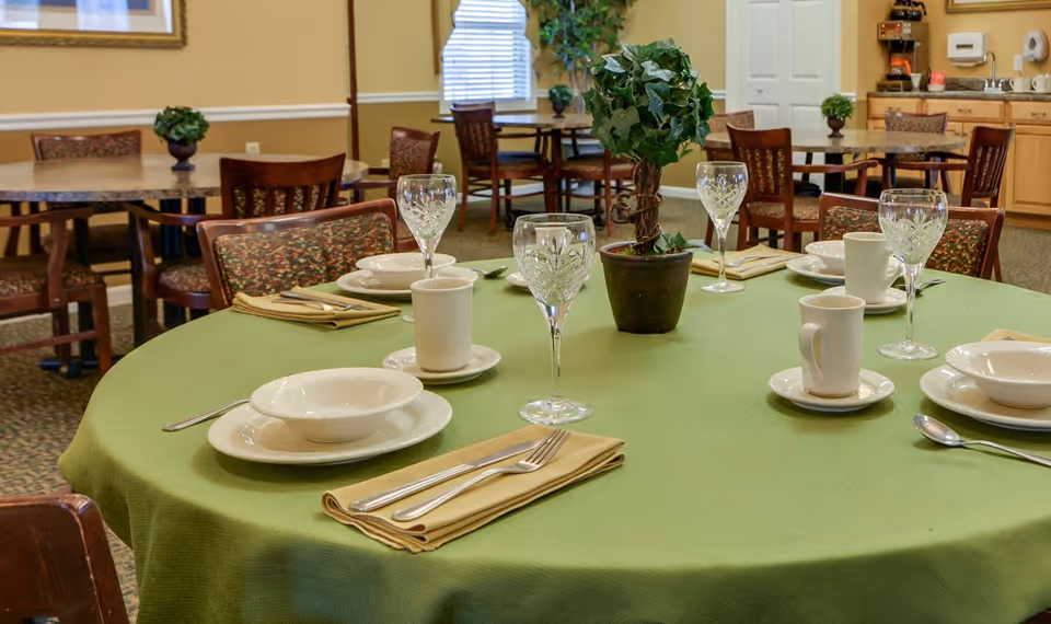 A dining room in a senior living facility with a round table covered by a green tablecloth set with white bowls, plates, cups, crystal wine glasses, and beige napkins with silverware. There is a small potted plant centerpiece on the table. In the background, there are more tables and chairs, a window with blinds, and a kitchenette area.