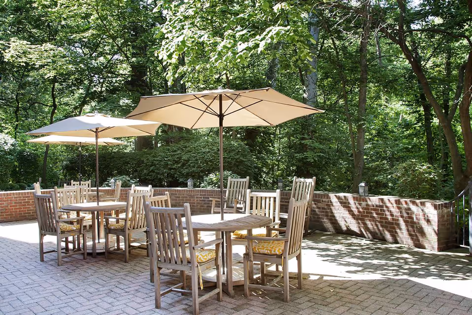 Outdoor patio area with round wooden tables and chairs, each table shaded by a large beige umbrella. The patio is paved with bricks and surrounded by a low brick wall, with dense green trees and foliage in the background.