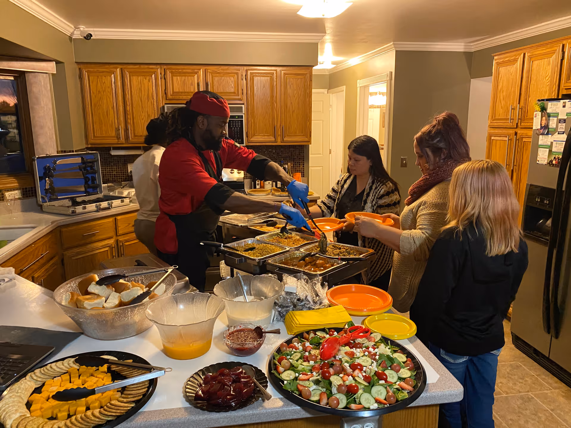 A kitchen scene with a chef serving food to three women standing in line with orange plates. The kitchen has wooden cabinets and a countertop filled with various dishes including salad, cheese and crackers, bread rolls, and other prepared foods.