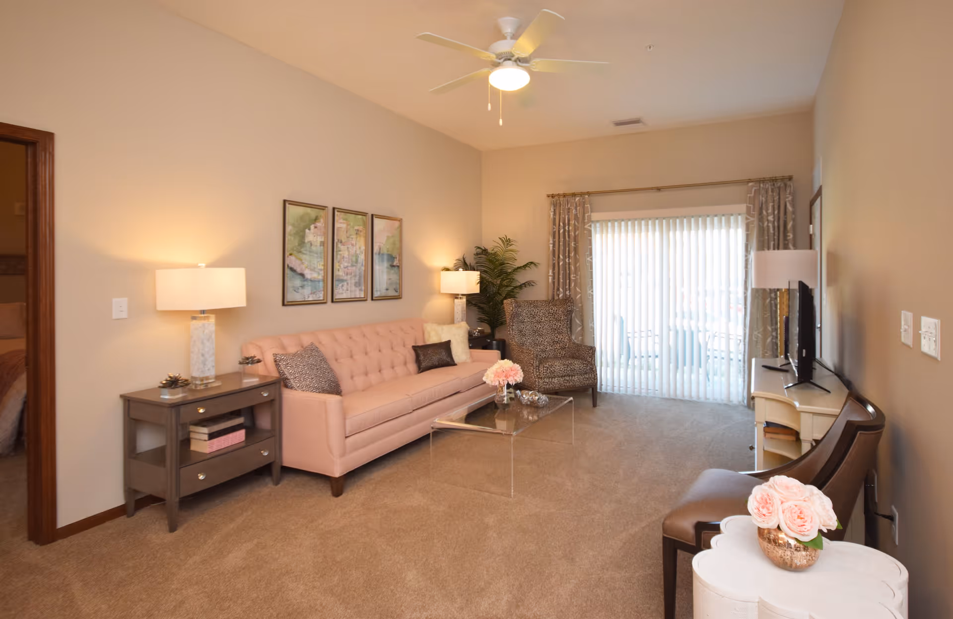 A cozy living room with a light pink tufted sofa, a patterned armchair, a glass coffee table with a flower arrangement, and a TV on a white stand. The room has beige walls, carpeted floor, two table lamps, a ceiling fan with light, and vertical blinds covering a sliding glass door. There is a small side table with a flower vase and a doorway leading to a bedroom.