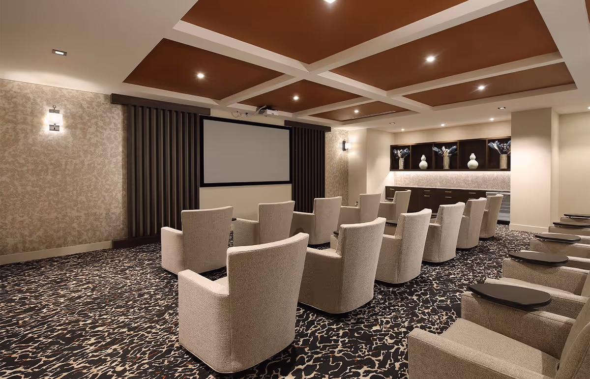 A modern theater room with multiple beige upholstered armchairs arranged in rows facing a large blank projection screen. The room features a patterned carpet, decorative wall sconces, a coffered ceiling with recessed lighting, and a back wall with built-in shelves displaying vases and floral arrangements.