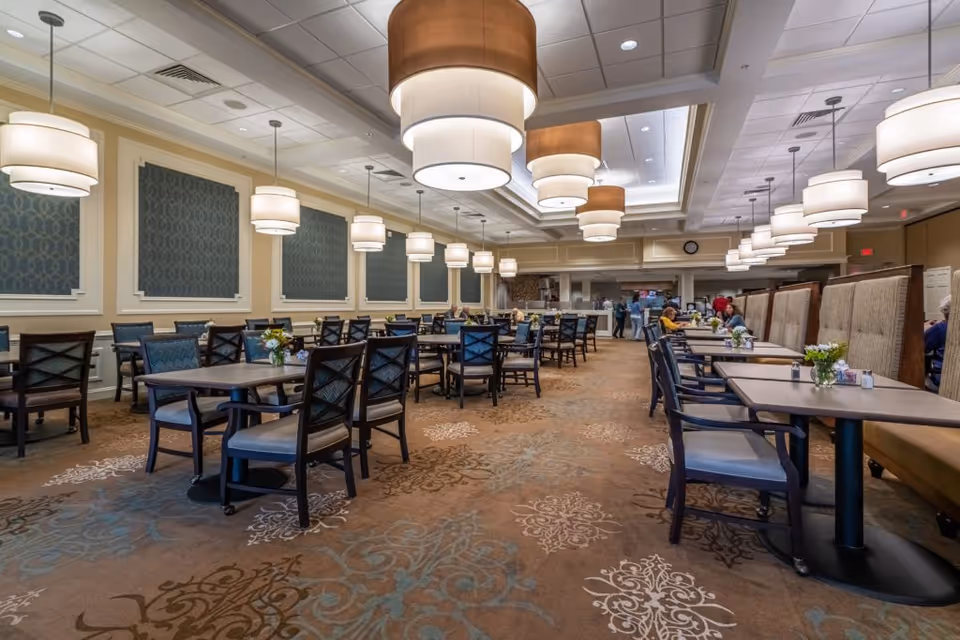 Spacious, well-lit dining room with multiple tables and chairs, booth seating along the right, and large pendant lights overhead.