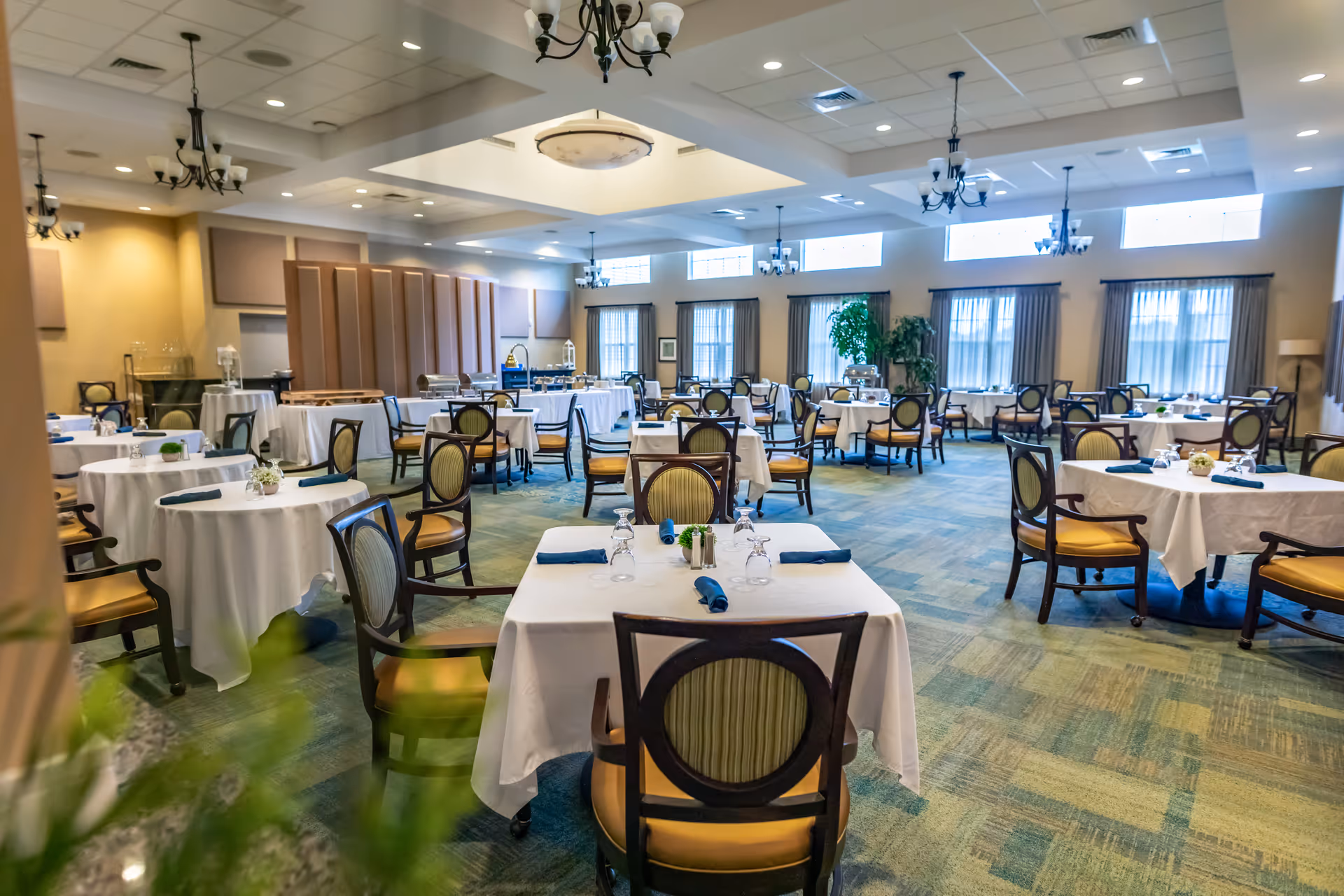 A spacious dining room with multiple round and square tables covered with white tablecloths, each set with glasses and napkins. The room has large windows with curtains, chandeliers hanging from the ceiling, and a carpeted floor with a green and blue pattern. There are wooden chairs with cushioned seats around the tables.