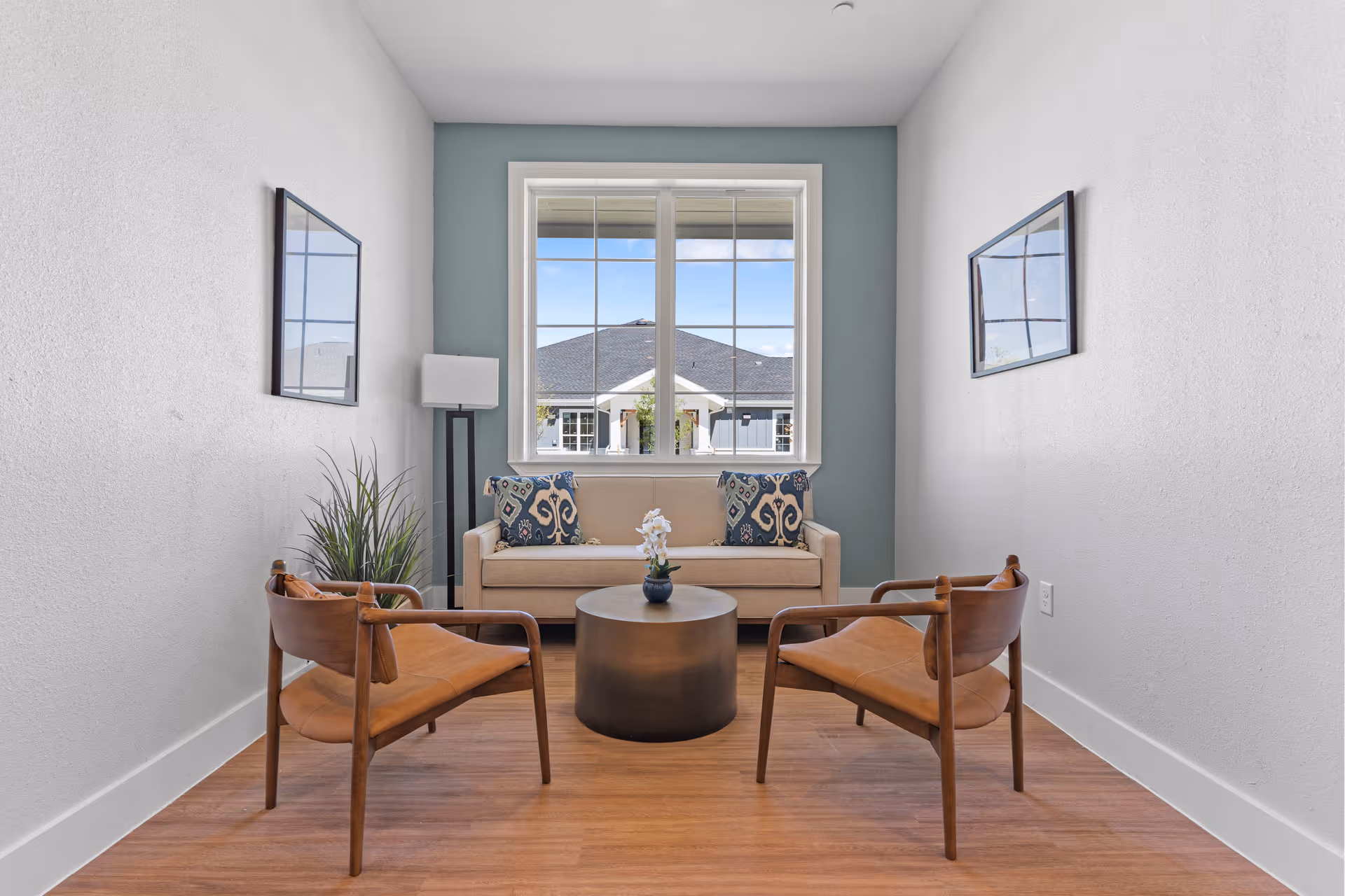 A small sitting area with a beige sofa adorned with two patterned cushions, two wooden chairs with brown leather seats, a round black coffee table with a small flower vase, a tall floor lamp, and two framed pictures on the white walls. A large window behind the sofa shows a view of a building with a dark roof and a blue sky with clouds.