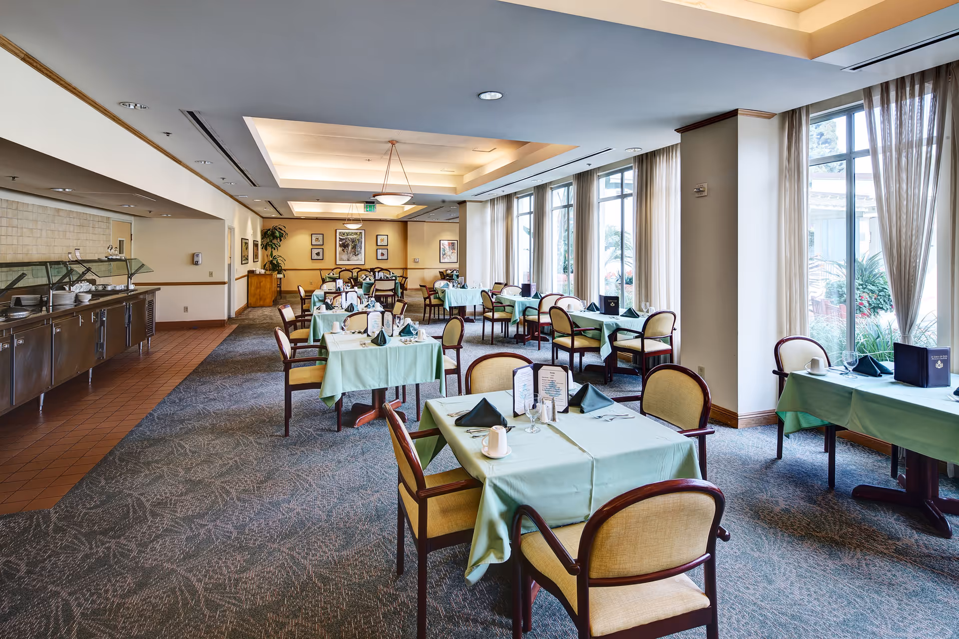 Bright dining room with multiple tables covered in green tablecloths and place settings, large windows with curtains, and a buffet counter along the left wall.