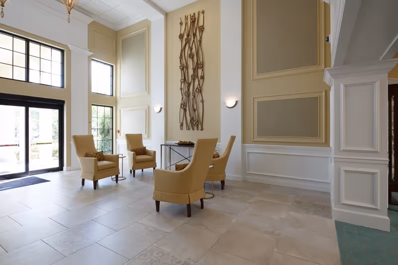 A bright and spacious seating area in a senior living facility with four beige armchairs arranged around a small round table. The room features large windows and glass doors letting in natural light, decorative wall paneling, and a tall metal wall sculpture. The floor is tiled in light colors, and there is a column with detailed molding on the right side.