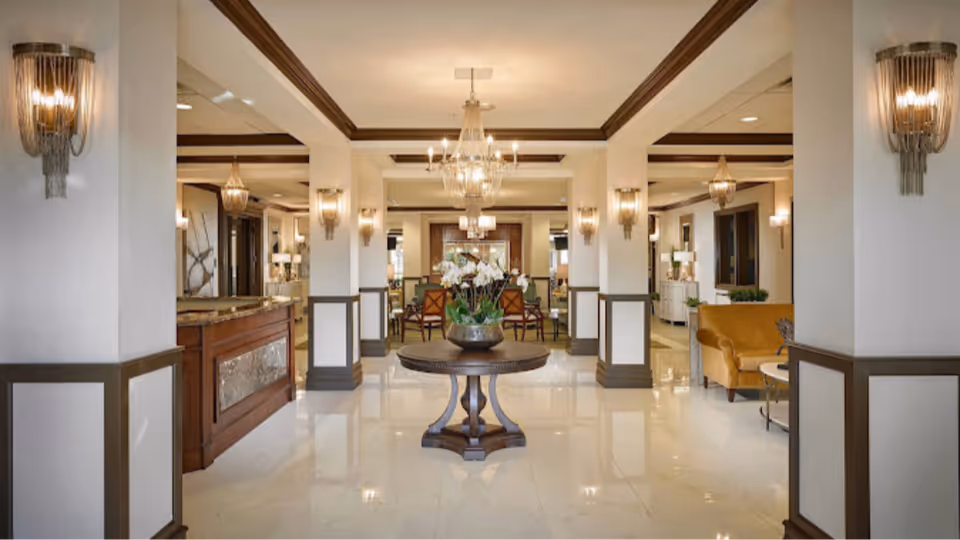 A spacious and elegant interior lobby area with polished marble floors, white walls with brown trim, and multiple chandeliers hanging from the ceiling. A round wooden table with a flower arrangement is centered in the foreground. There are seating areas with chairs and sofas visible in the background.