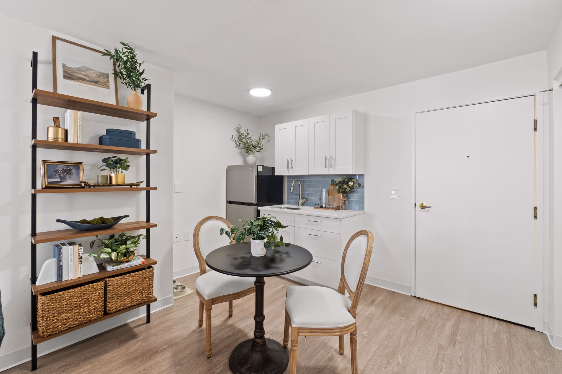 Small airy kitchenette and dining nook with a round table and two chairs, open shelving, stainless fridge, and white cabinets.