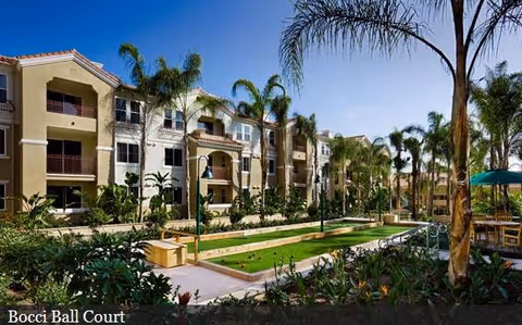 Outdoor bocce ball court surrounded by palm trees and landscaped greenery with a multi-story residential building in the background under a clear blue sky.