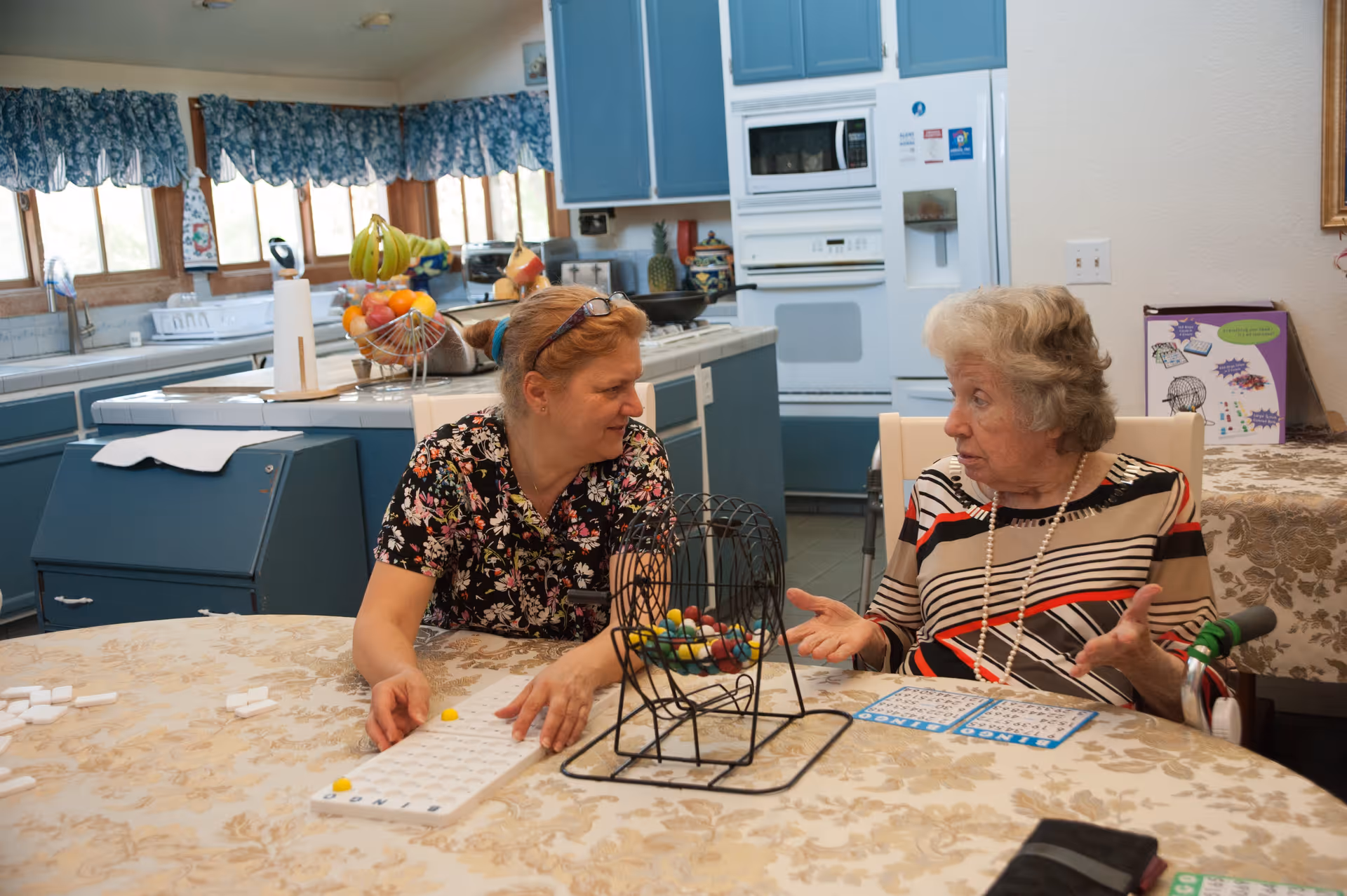 Two women sitting at a table in a kitchen playing a game with a bingo cage and bingo cards. The kitchen has blue cabinets, a white oven, and a refrigerator. The table is covered with a floral tablecloth and there is a fruit basket on the kitchen counter.