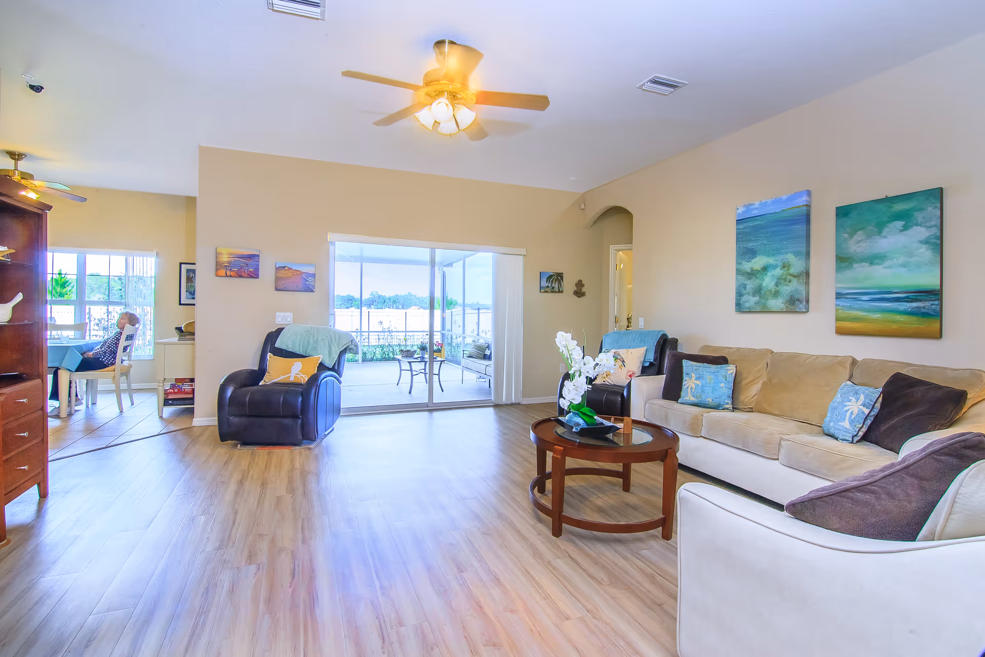 Bright and spacious living room with light wood flooring, beige walls, and ceiling fans. The room features a beige sofa with blue and brown cushions, a round wooden coffee table with a white orchid centerpiece, and a black recliner chair with a green throw and orange pillow. Two ocean-themed paintings hang on the wall above the sofa. Sliding glass doors lead to a screened-in porch with outdoor seating and a view of greenery and water. In the background, a dining area with a person sitting at a table is partially visible.