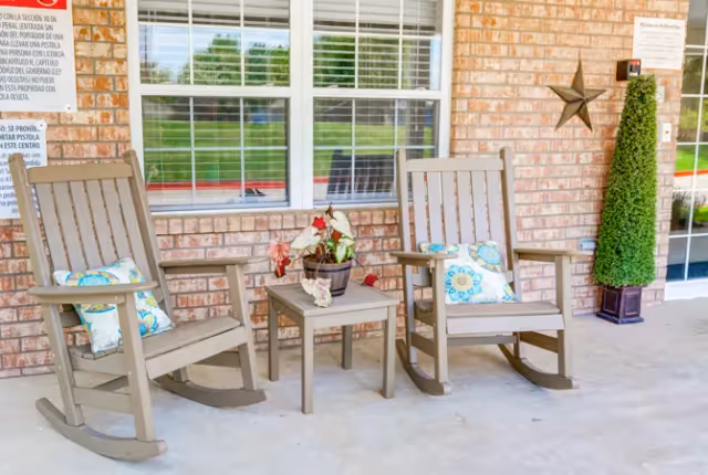 Two beige wooden rocking chairs with colorful floral cushions on a covered porch in front of a brick wall with a window. Between the chairs is a small matching table with a potted plant. A tall, cone-shaped topiary in a planter and a decorative star are mounted on the brick wall near a glass door.