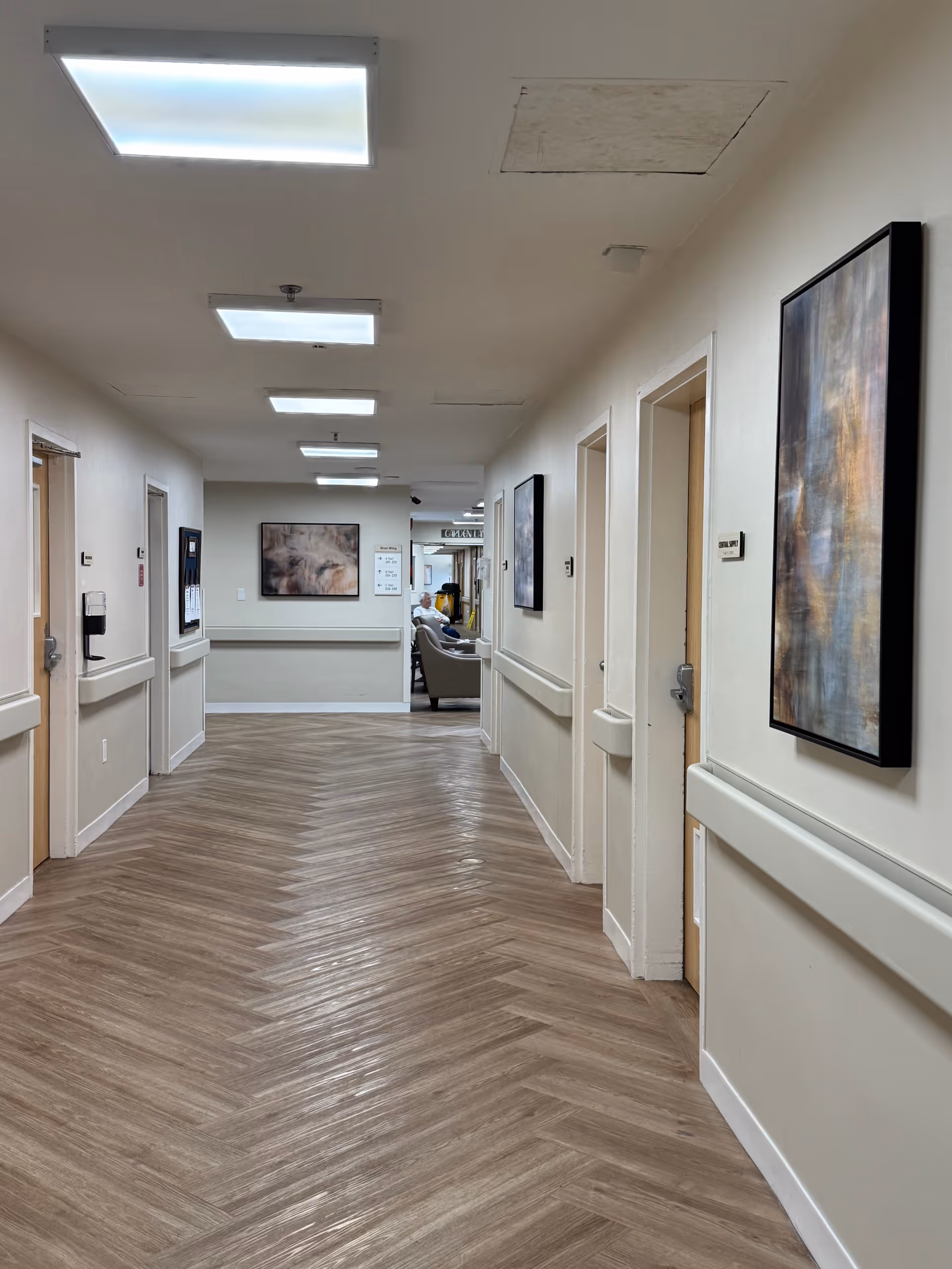 A well-lit interior hallway in a care facility with herringbone wood-patterned flooring, handrails, doors, and framed artwork leading to a seating area.