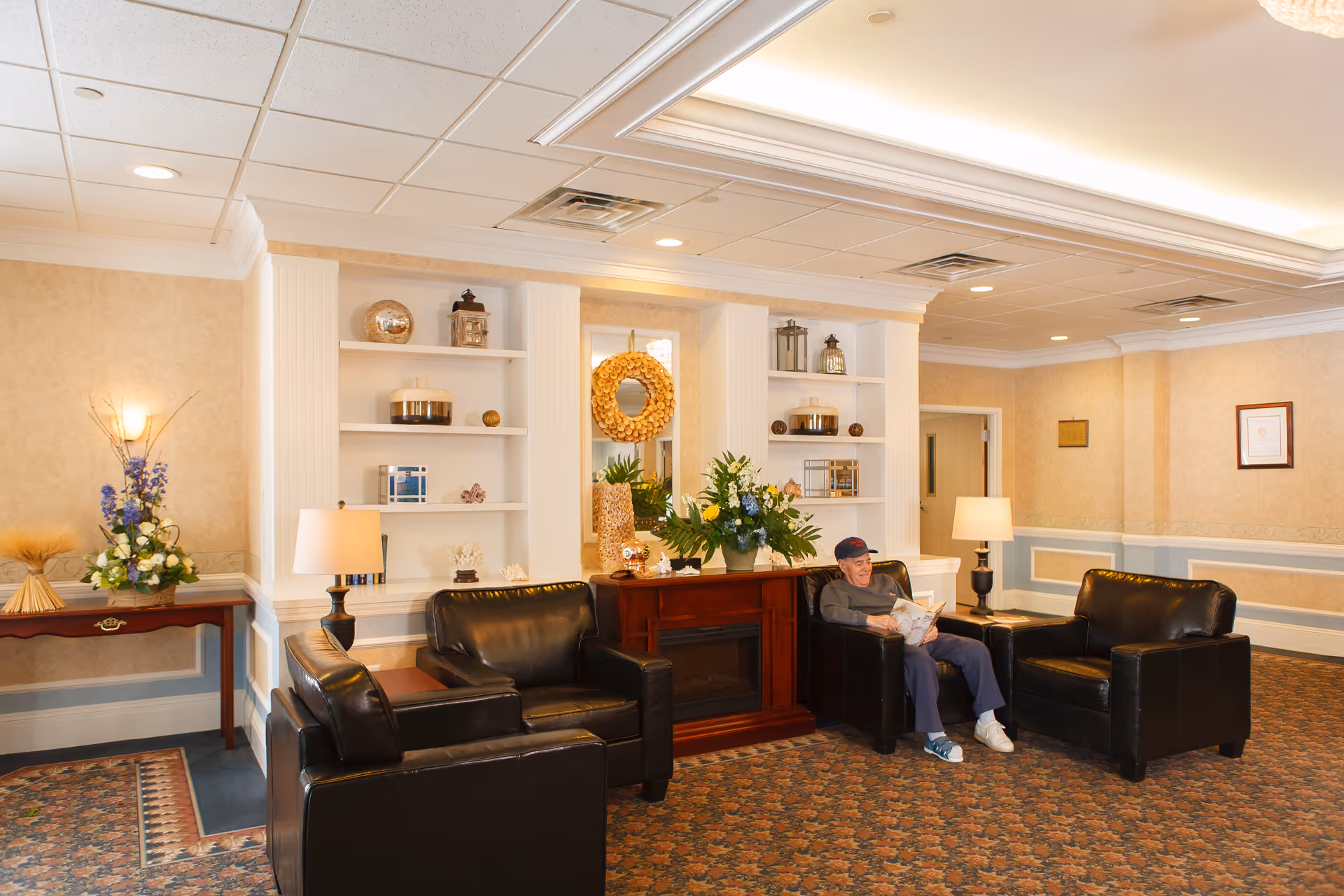 A cozy sitting area in a senior living facility with three black leather armchairs arranged around a wooden electric fireplace. A man wearing a cap is seated in one of the chairs reading a newspaper. The room has beige walls, built-in white shelves with decorative items, two table lamps, floral arrangements, and a patterned carpet.
