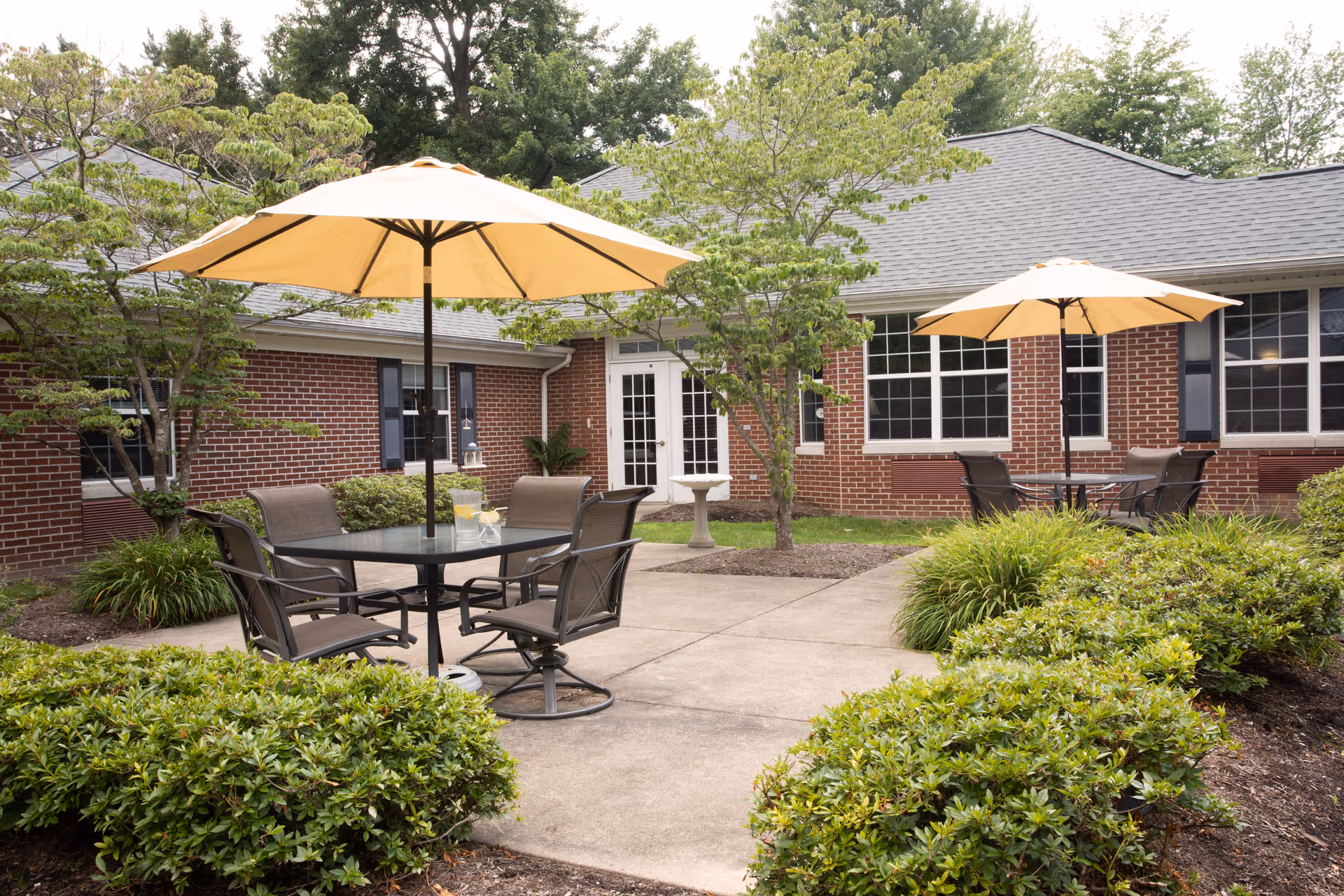 Outdoor patio area at Governor's Pointe with two round glass tables, each shaded by a large beige umbrella. Each table is surrounded by four chairs. The patio is surrounded by green bushes, small trees, and a brick building with multiple windows and a white door in the background.